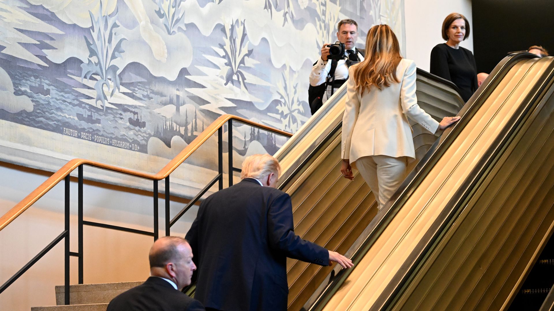 The backs of President Trump, wearing a long, navy jacket, and first lady Melania Trump, wearing a cream suit, walking up steps beside an escalator.
