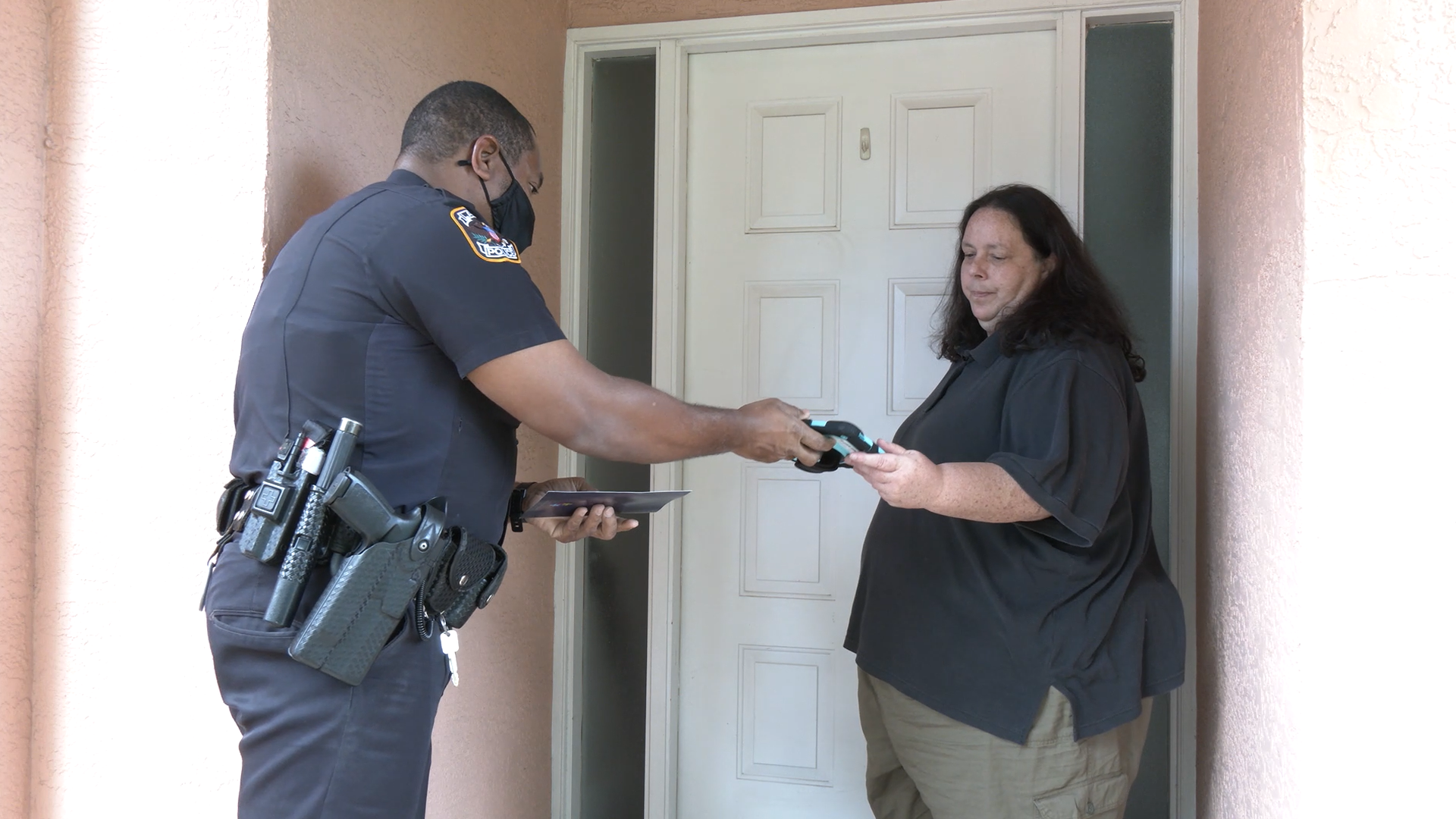 An officer of the law hands a civilian a tablet before the door of the civilian's home.