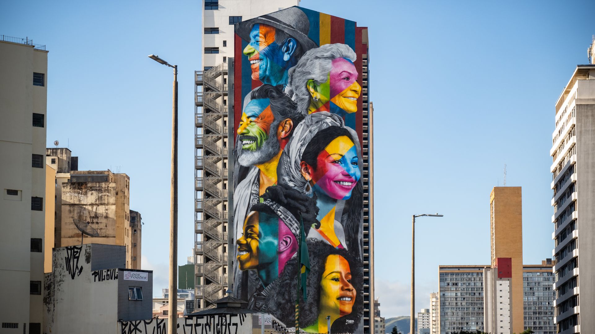 A multicolored mural of people's faces on a building with a blue sky in background.