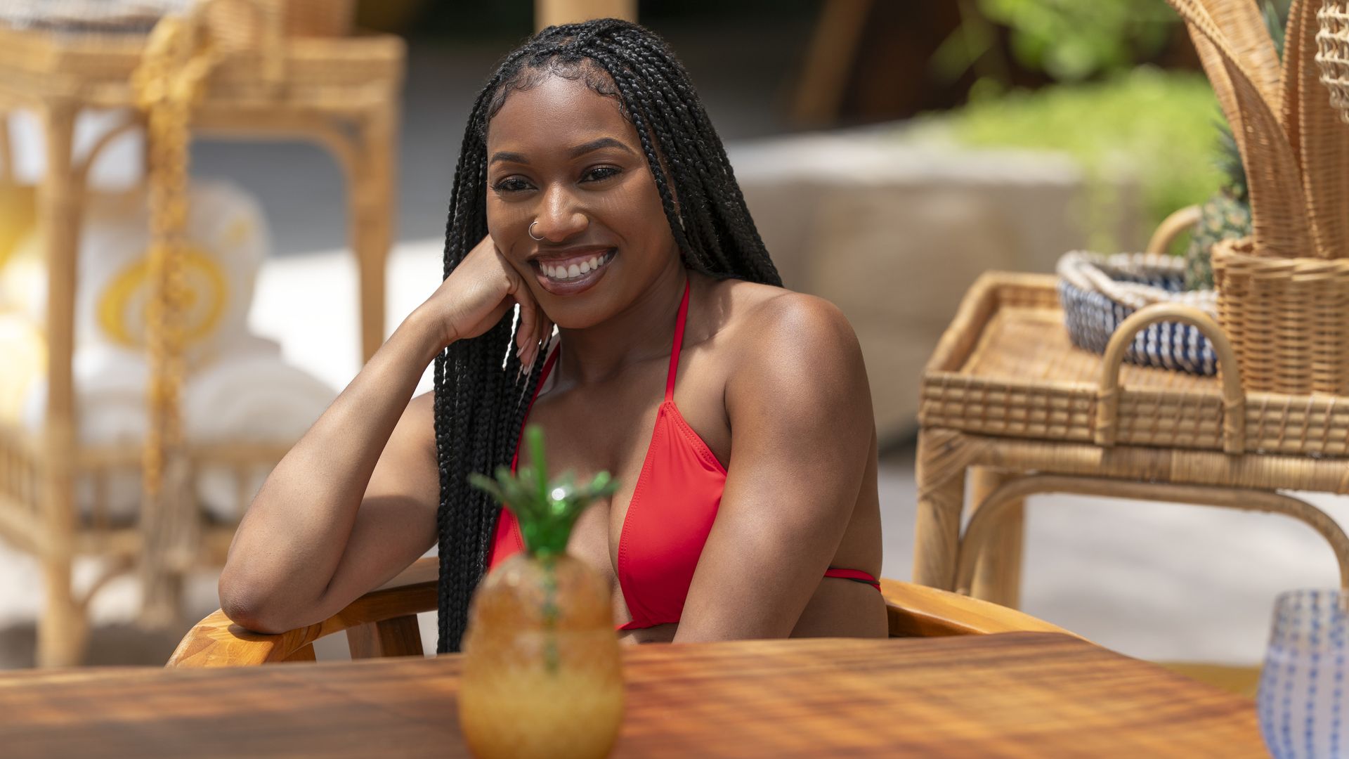 Smiling woman with long braids and nose ring, wearing a red bikini top, sitting at wooden table with a pineapple-shaped drink in front of her in a sunny, wicker-furnished outdoor setting.