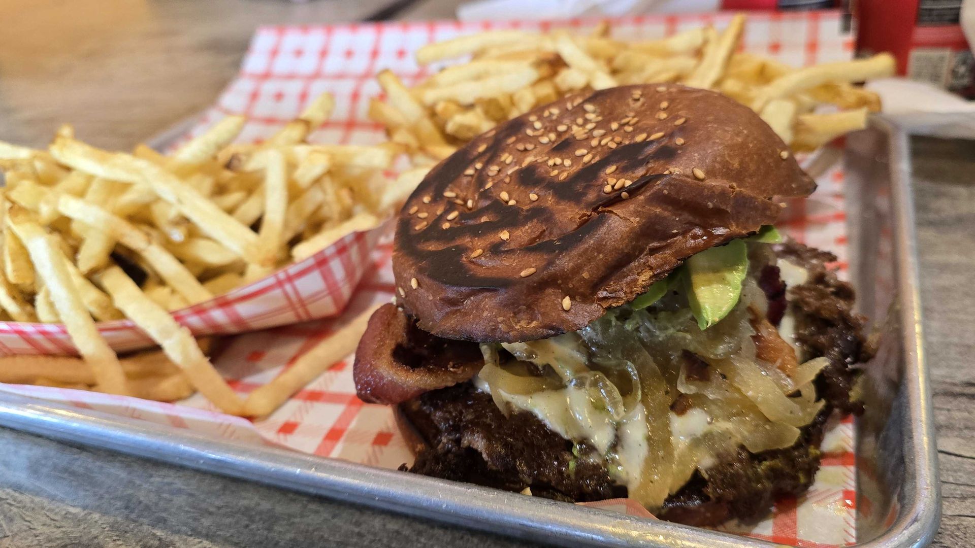 Cheeseburger with sesame bun, melted cheese and onions on a metal tray, beside a heap of French fries in a red-checkered basket on a wooden table.