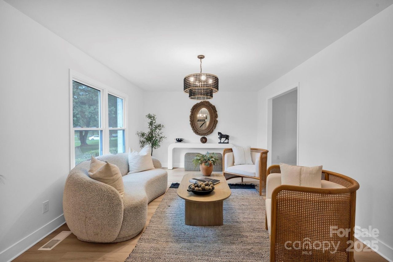 Modern living room with curved beige sofa, two wooden armchairs with white cushions, oval wooden coffee table, decorative plants, black chandelier, and large windows showing outdoor greenery.