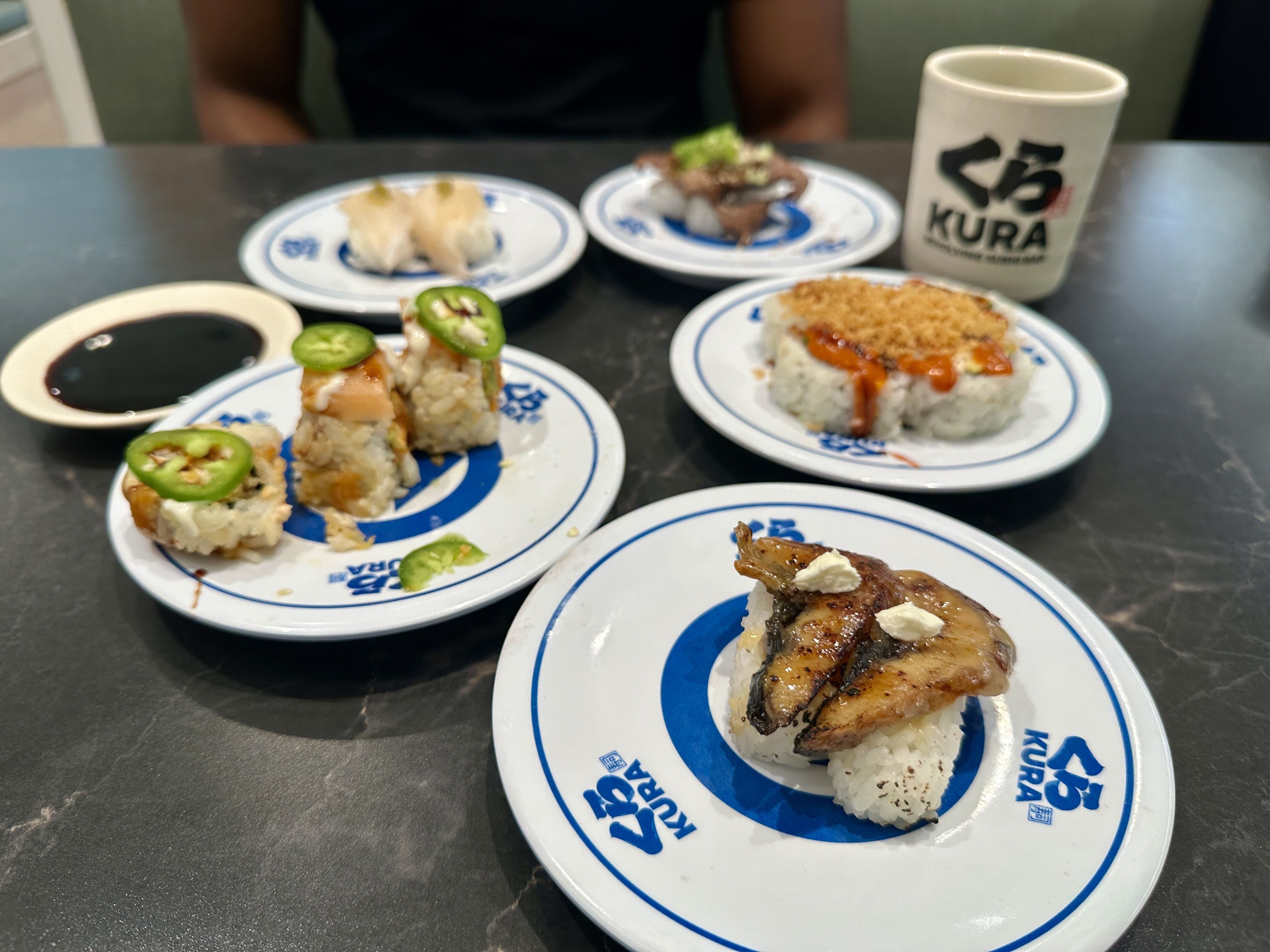 Assorted sushi plates on a dark table, topped with jalapeño slices and sauces; a white mug with the "KURA" logo sits in the background as a person is blurred behind the table.