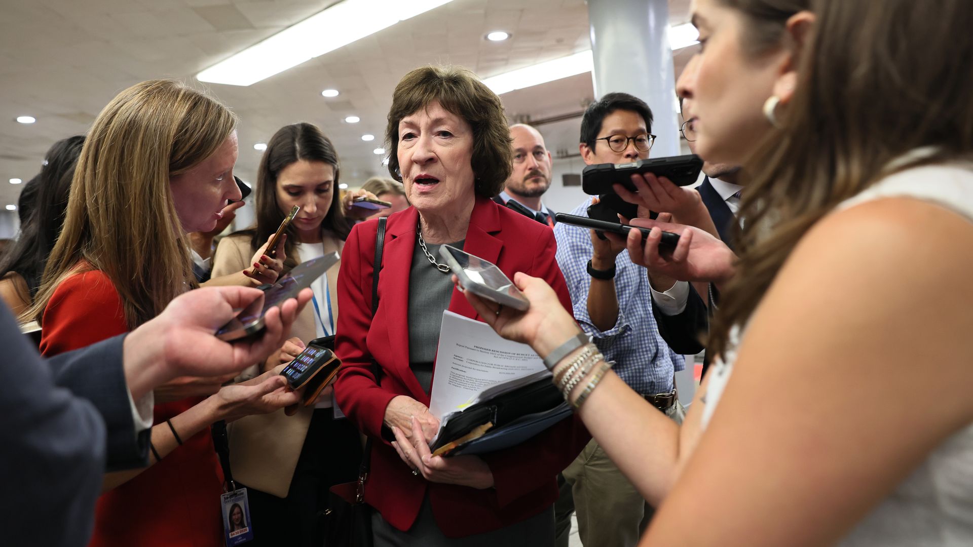 Sen. Susan Collins, wearing a red blazer and holding a bill and folders, speaks to reporters in a white, windowless basement.