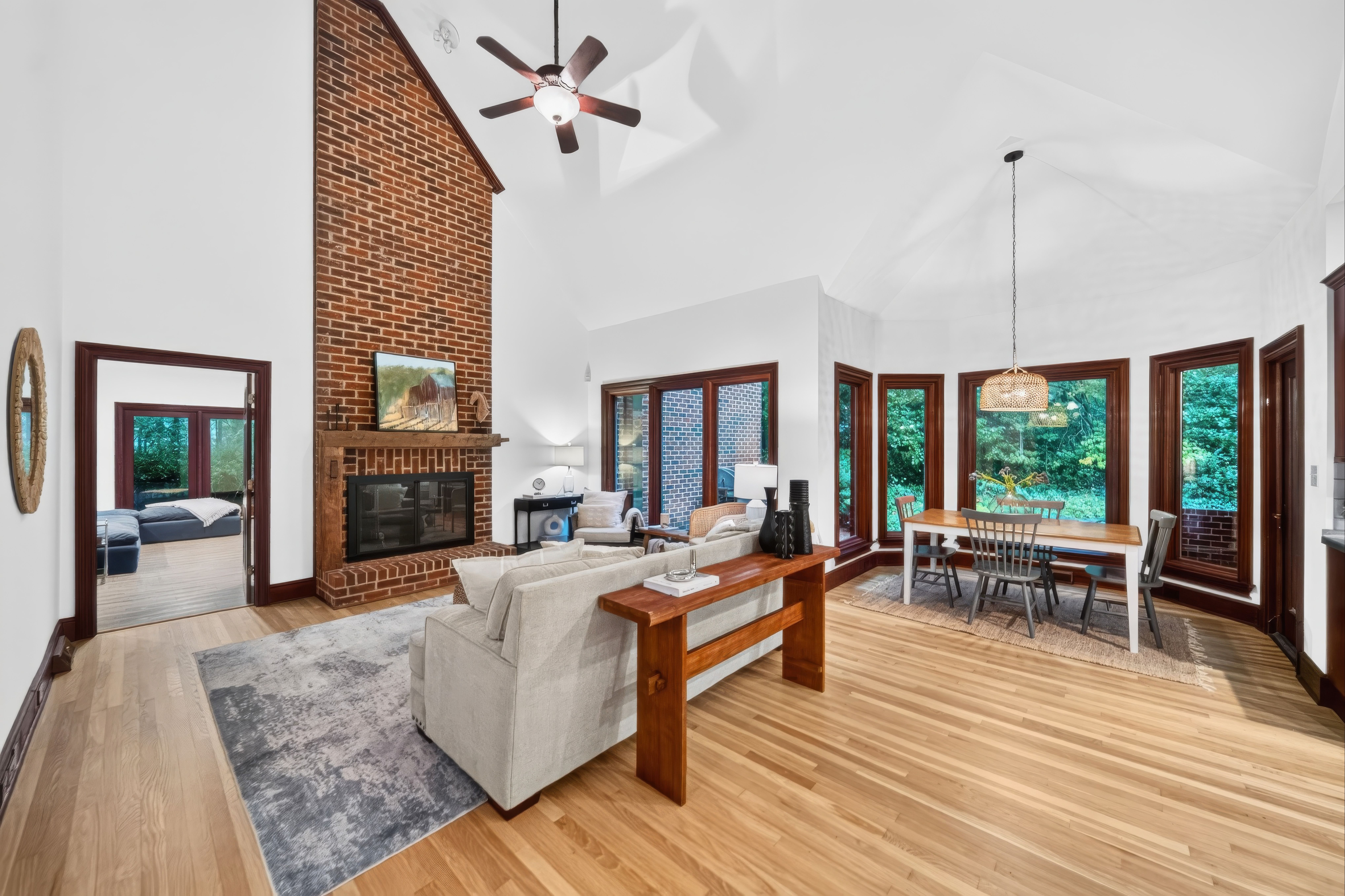 Bright open living area with a brick fireplace, ceiling fan, and tall white ceilings. Large wooden-framed windows, gray sofas, a wooden coffee table, and a dining table with wicker pendant light.