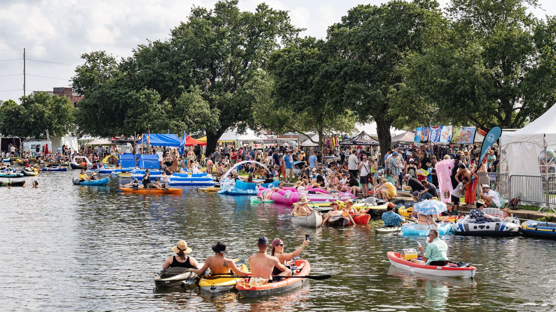 People in kayaks fill a crowded bayou next to a crowded stretch of land with vendor tents.
