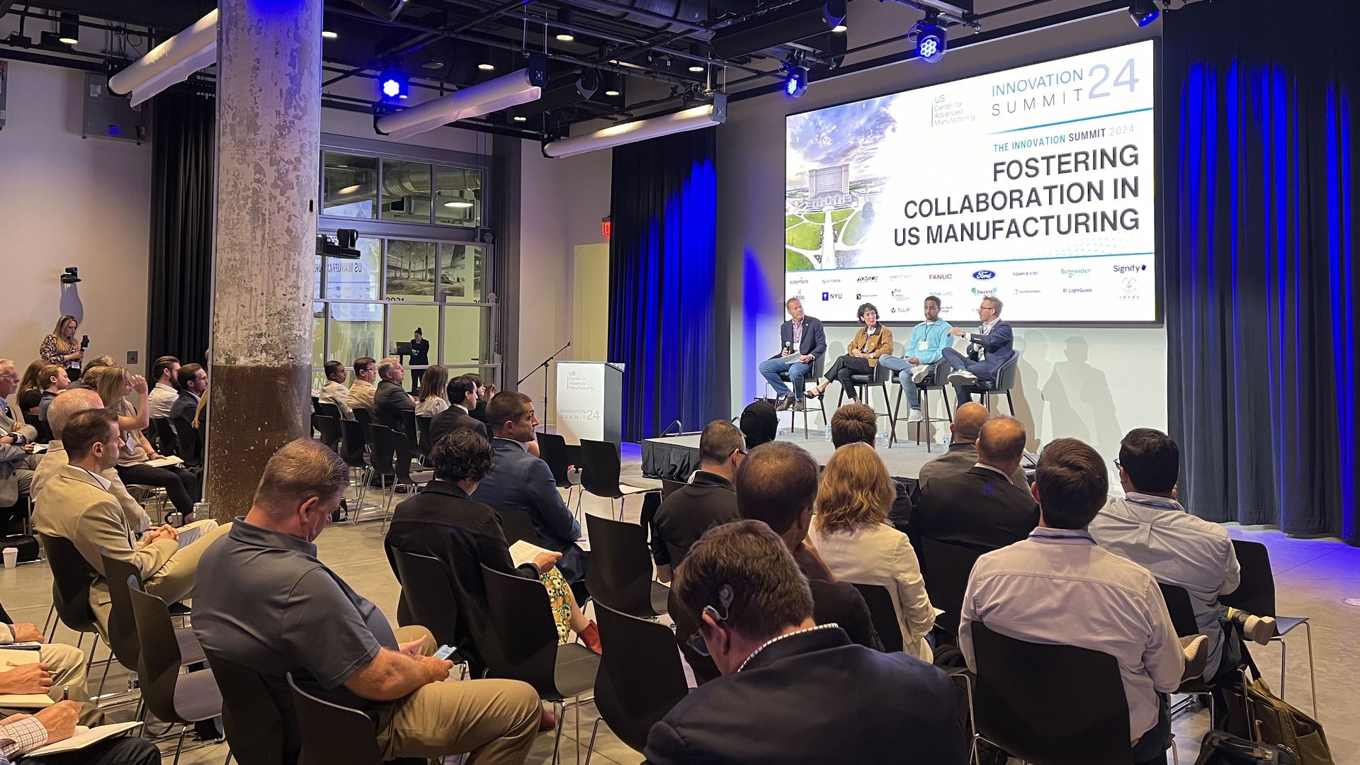 A crowd looks on at a panel with a presentation behind them titled "fostering collaboration in U.S. manufacturing."