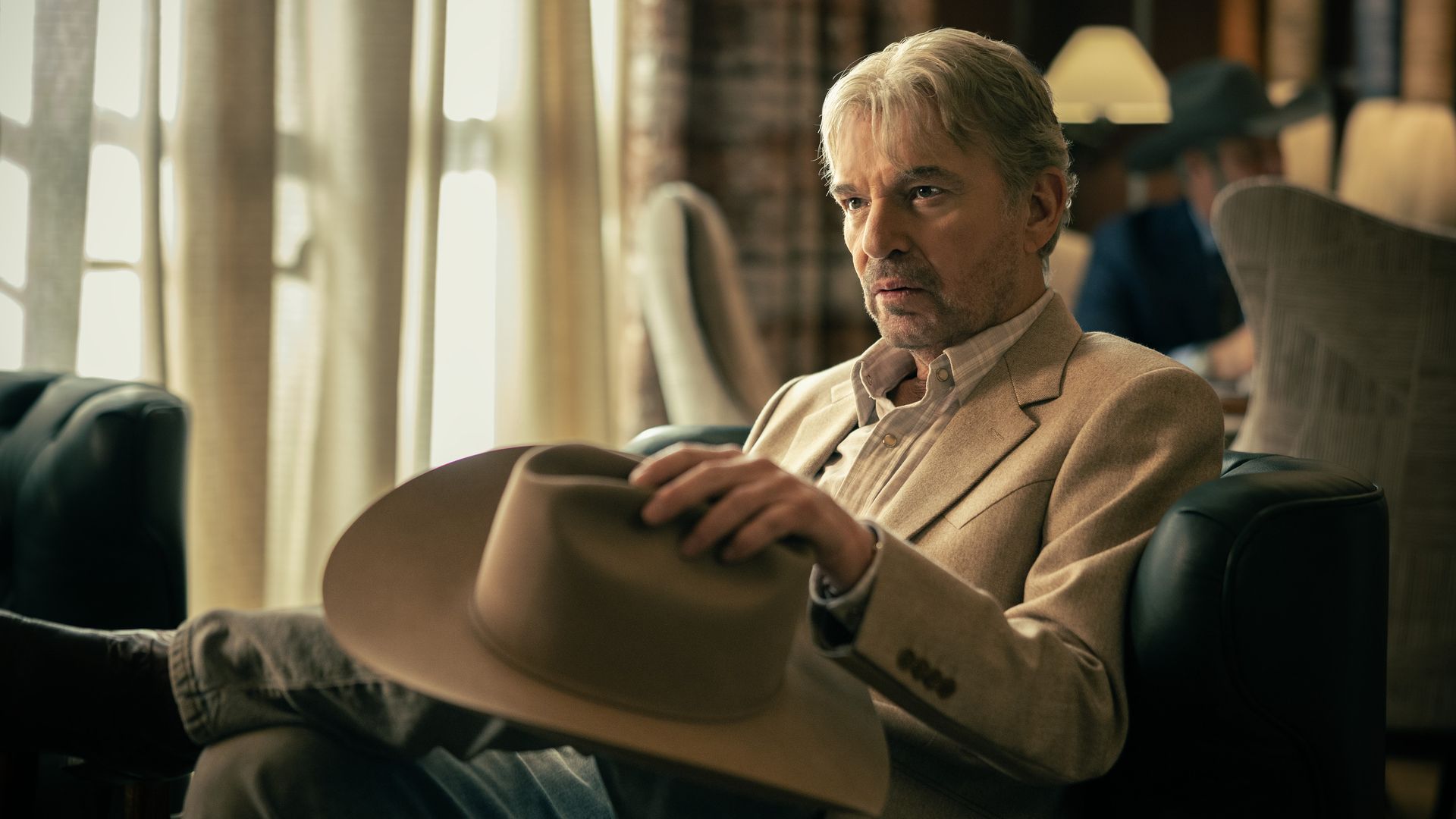 Older man with gray hair sitting indoors in a beige blazer, holding a tan cowboy hat, with light curtains and another person wearing a dark hat blurred in the background.