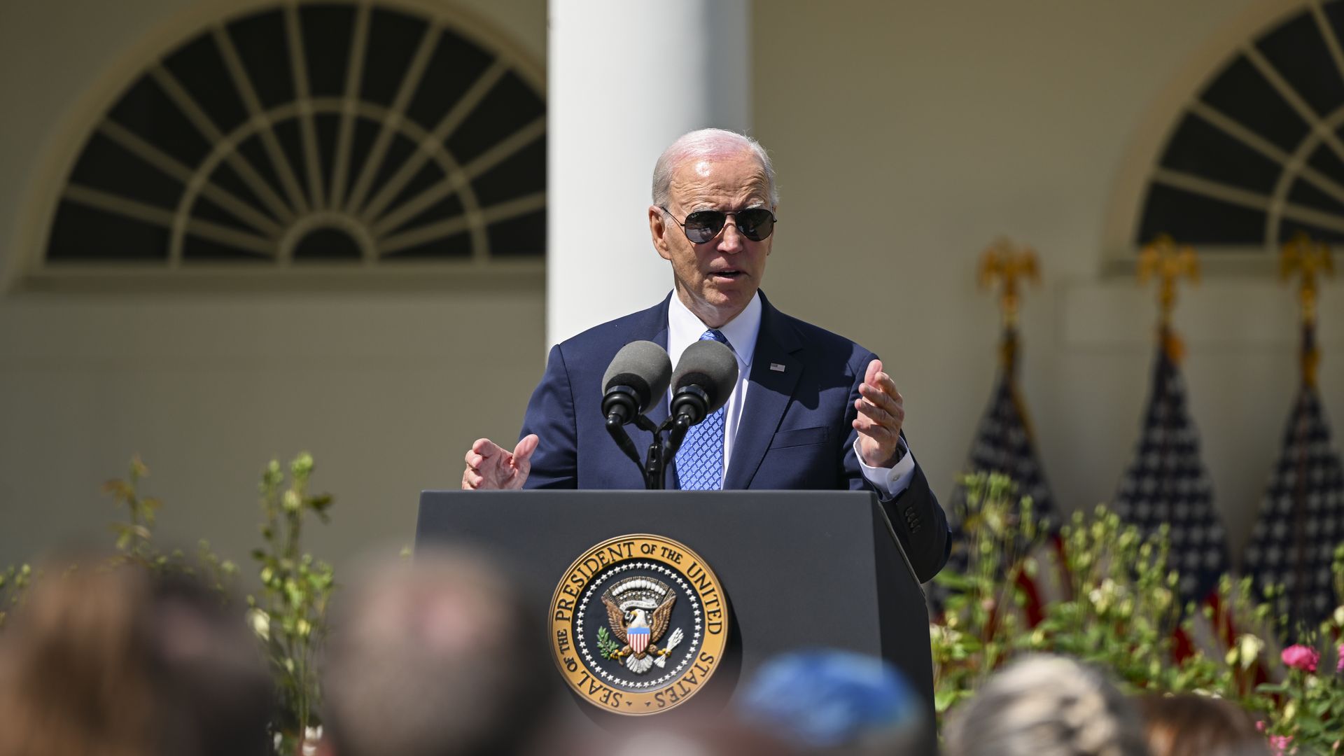President Joe Biden, wearing a blue suit, white shirt, blue tie and aviator sunglasses, speaks to a crowd behind a podium in the White House rose garden.