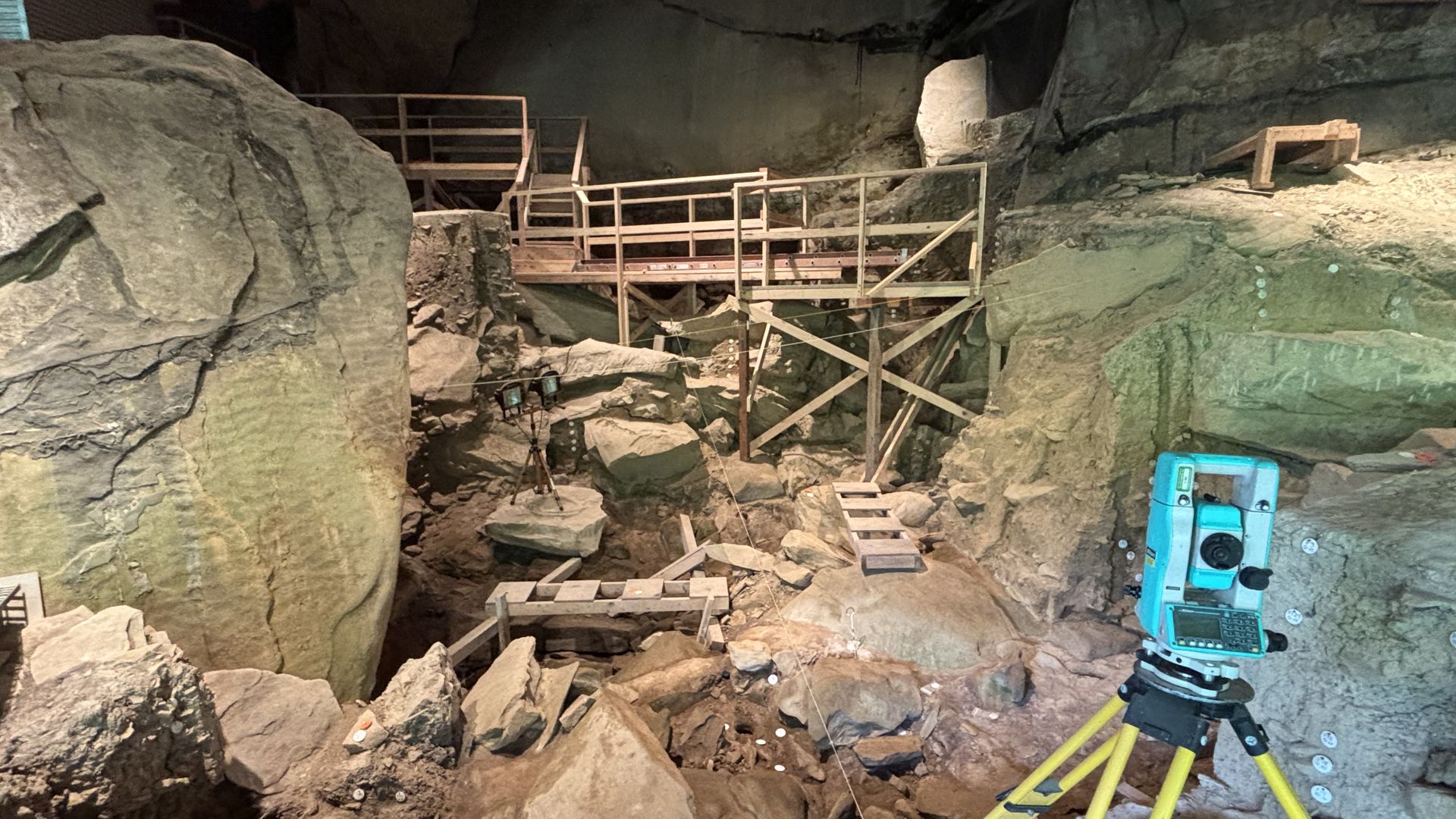 Inside a rocky cave, wooden scaffolding and stairs form a walkway over rubble. A teal surveying instrument rests on yellow cables to the right, with rough rock walls overhead.