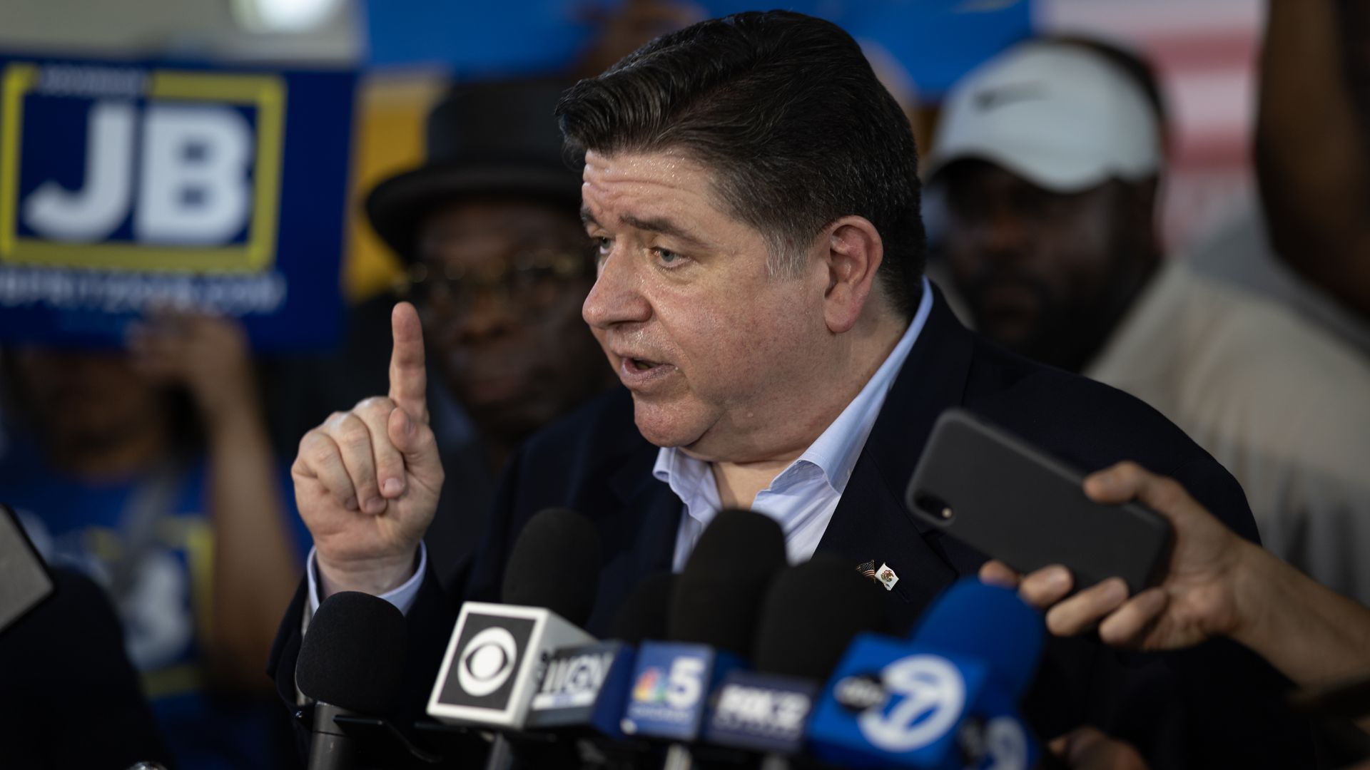 Photo of a man at a podium with his finger raised with microphones in his face. 