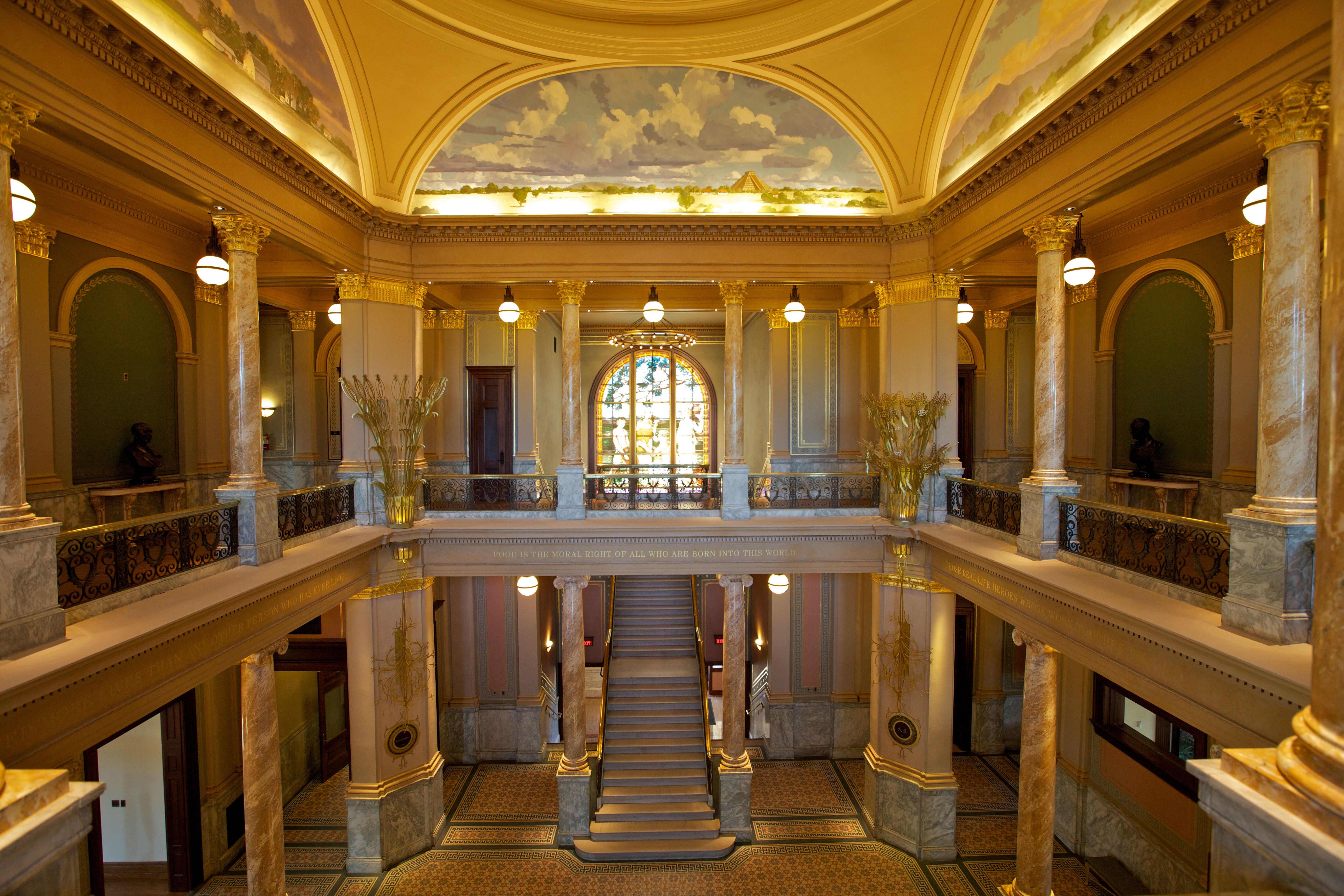 Grand neoclassical atrium with marble columns and balconies, a central staircase, and a painted ceiling of clouds over a distant pyramid; warm globe lights illuminate gold-trimmed arches.