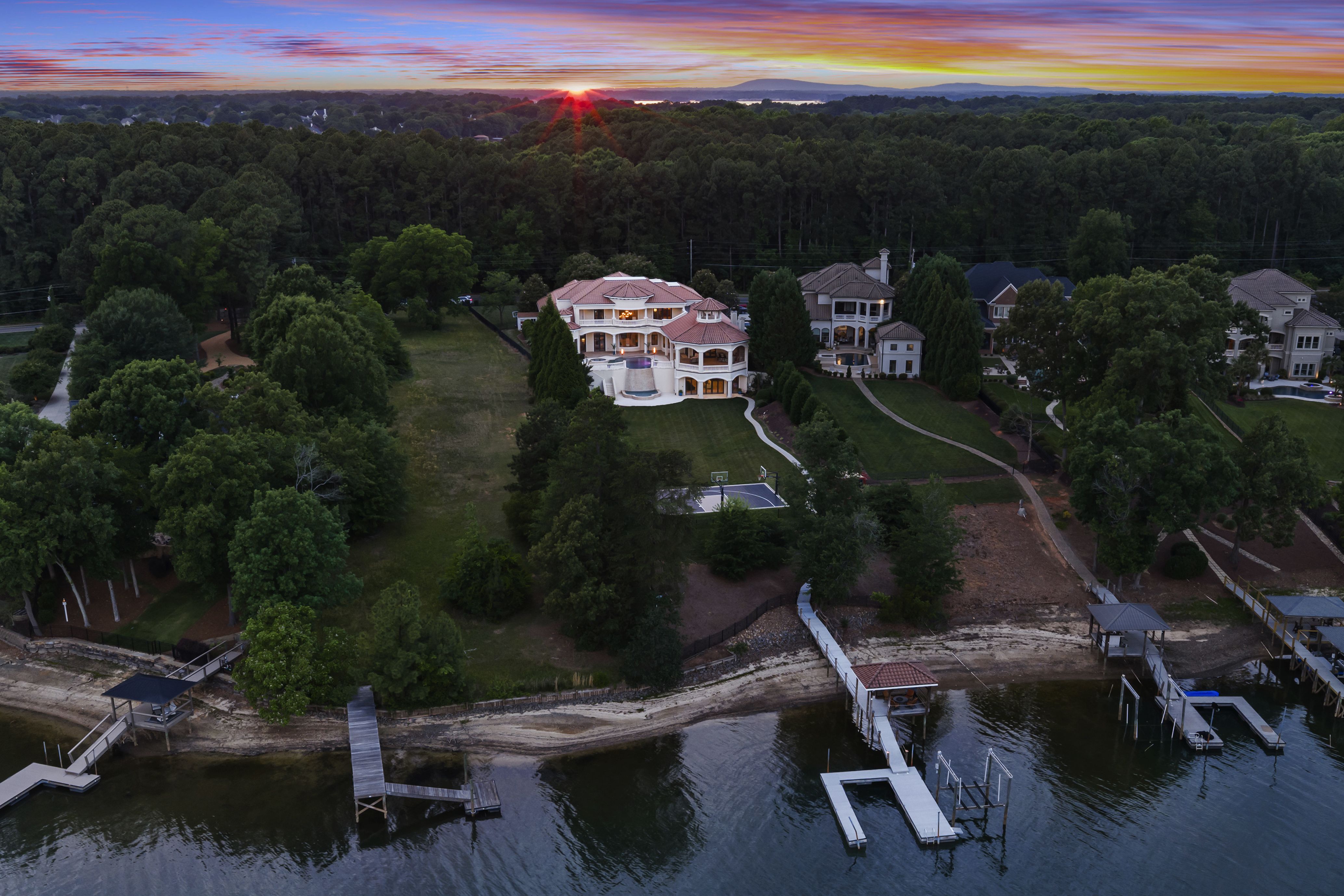 Aerial view of a white mansion with a red-tiled roof on a lakeside lot, surrounded by dense trees and nearby houses. Wooden docks extend into calm water as an orange-pink sunset fills the sky.