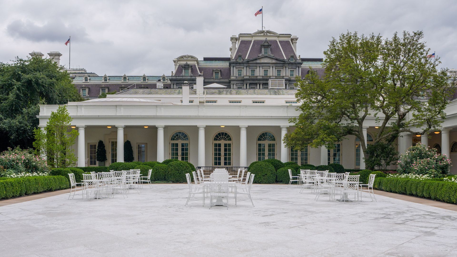 White patio with empty white metal chairs and tables, surrounded by green bushes and trees, in front of a white columned building with arched windows under a cloudy sky.