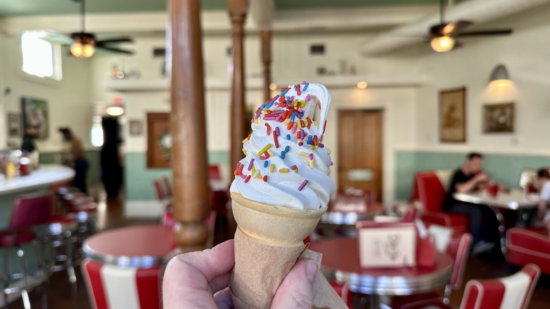 A vanilla soft-serve cone with colorful sprinkles held in a hand, in a retro diner with red-and-white seating, wood columns, and ceiling fans in the background.