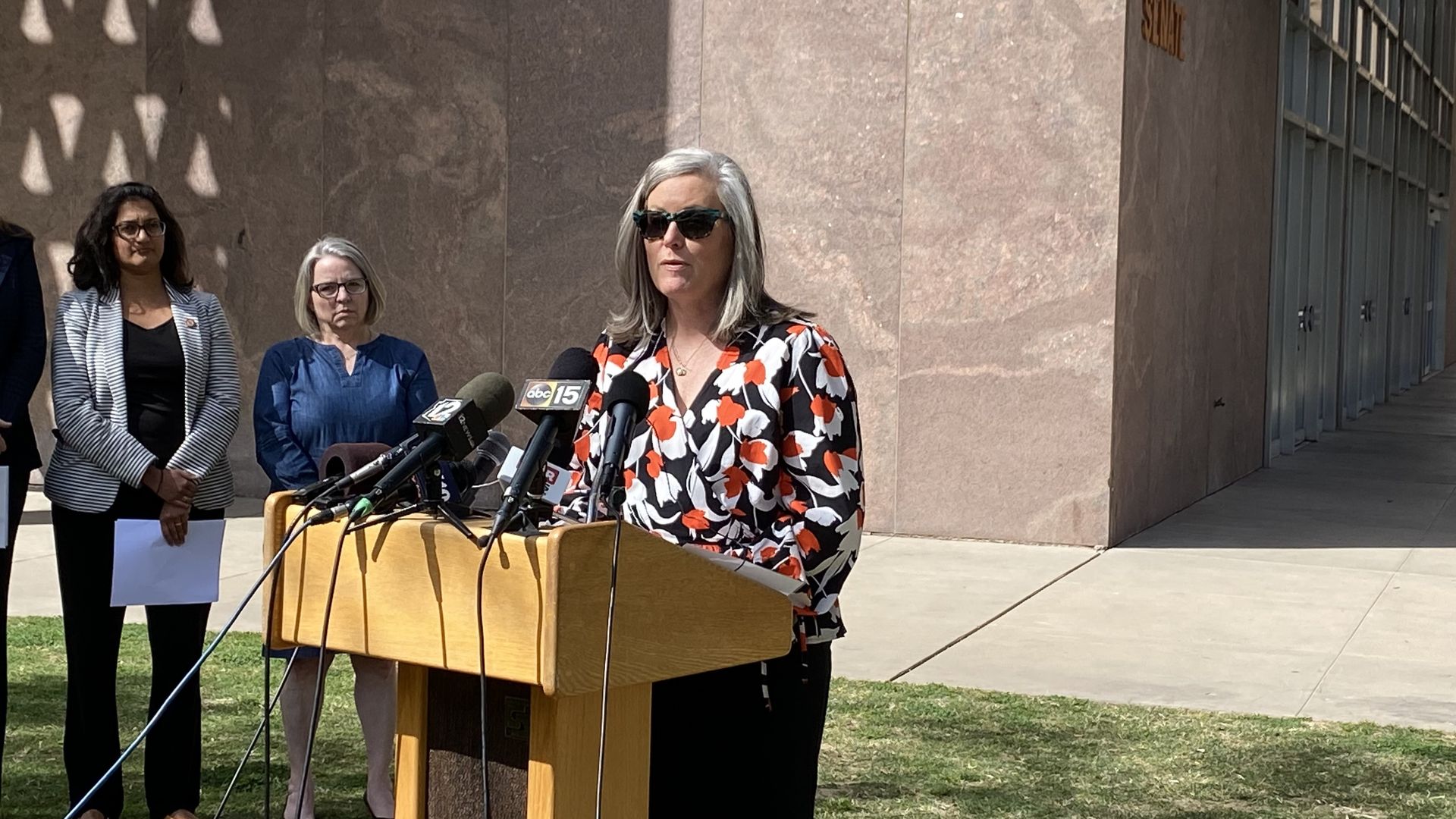 A woman speaks outdoors at a lectern with several microphones on it.