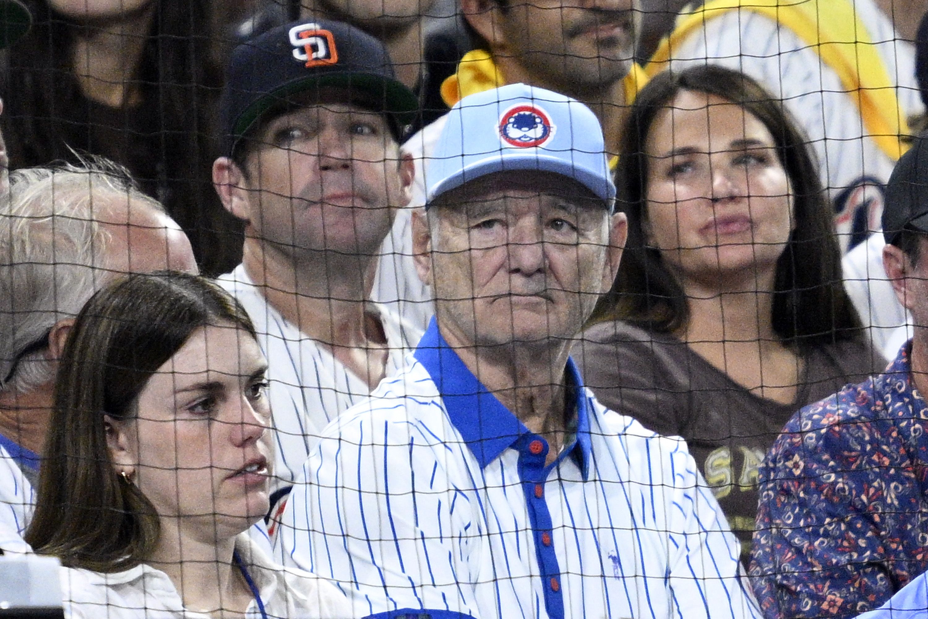 Crowd of spectators behind protective netting at a baseball stadium; a man in a light-blue cap with a red-and-white circular logo and blue-striped shirt sits near others.