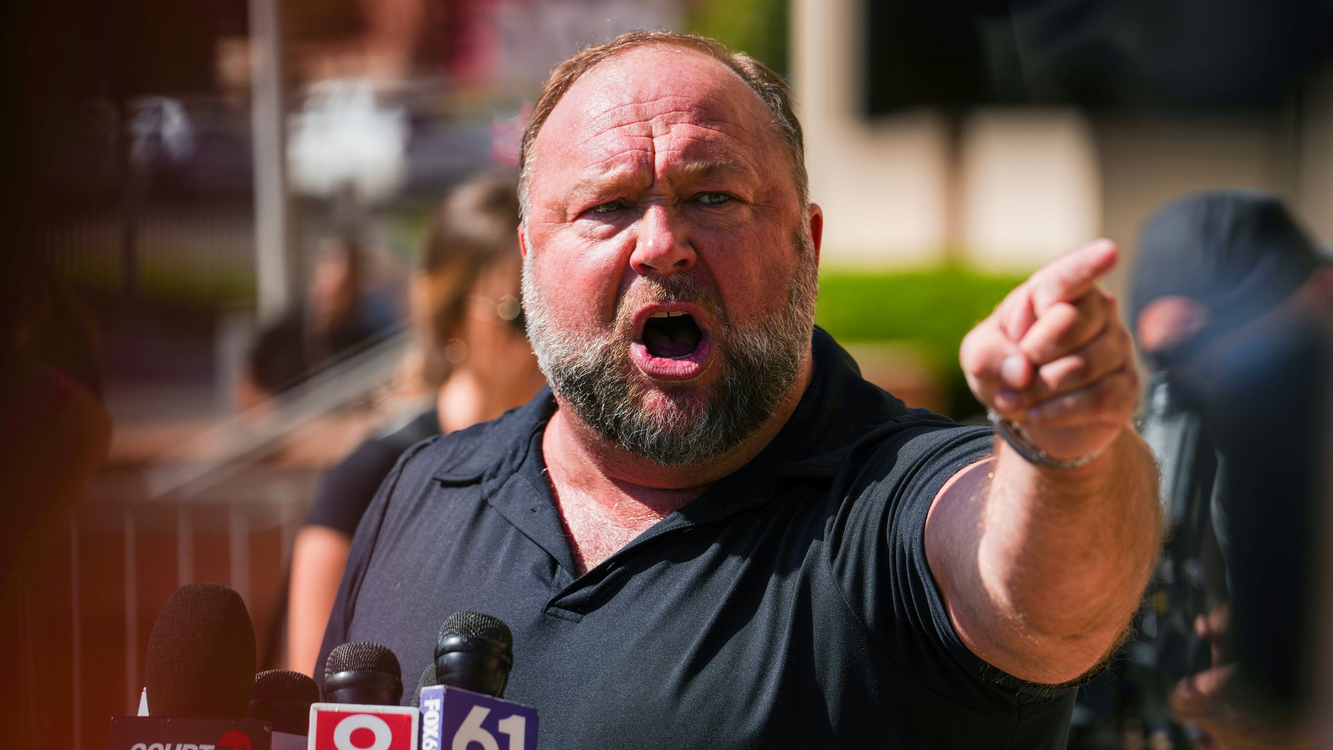 Alex Jones, wearing a black shirt with a collar and short sleeves, points while speaking into microphones.