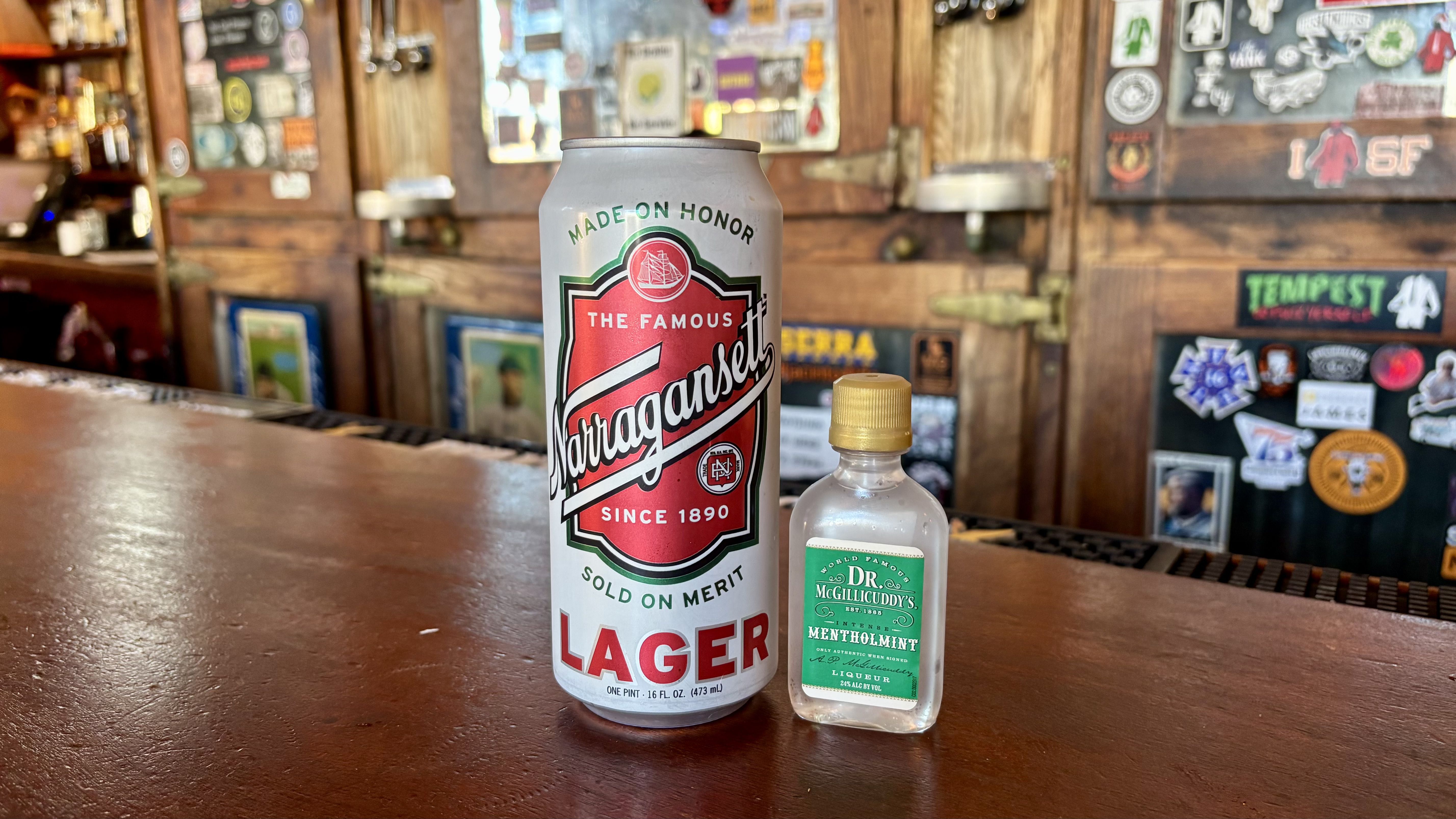 A large can of Narragansett Lager beer next to a small bottle of Dr. McGillicuddy's Mentholmint liquor on a wooden bar counter with stickers and posters in the background.