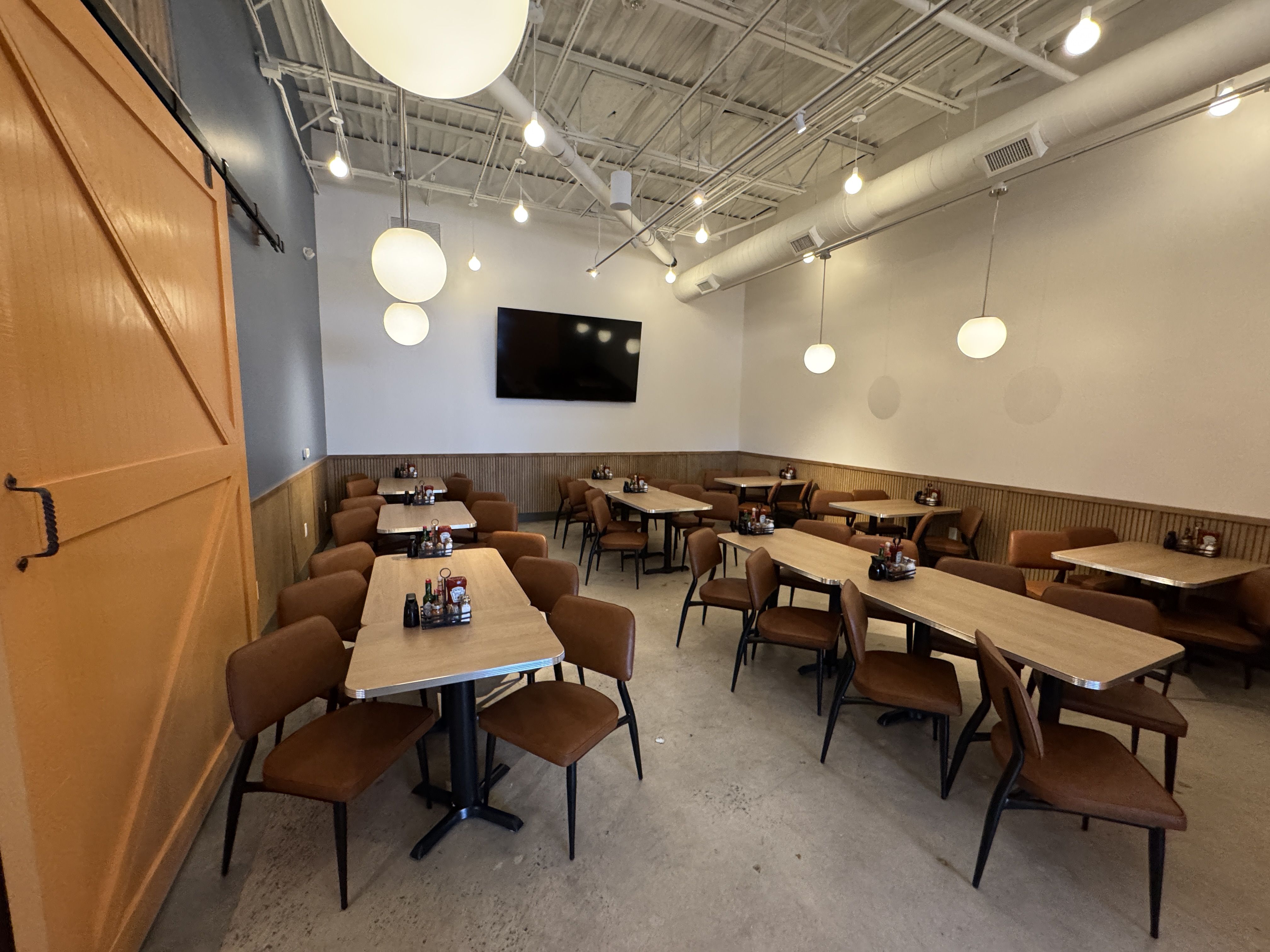 Interior of a modern restaurant with wood tables, brown chairs, pendant lights, an orange sliding barn door on the left, white walls with wood paneling, and a large TV on the back wall.