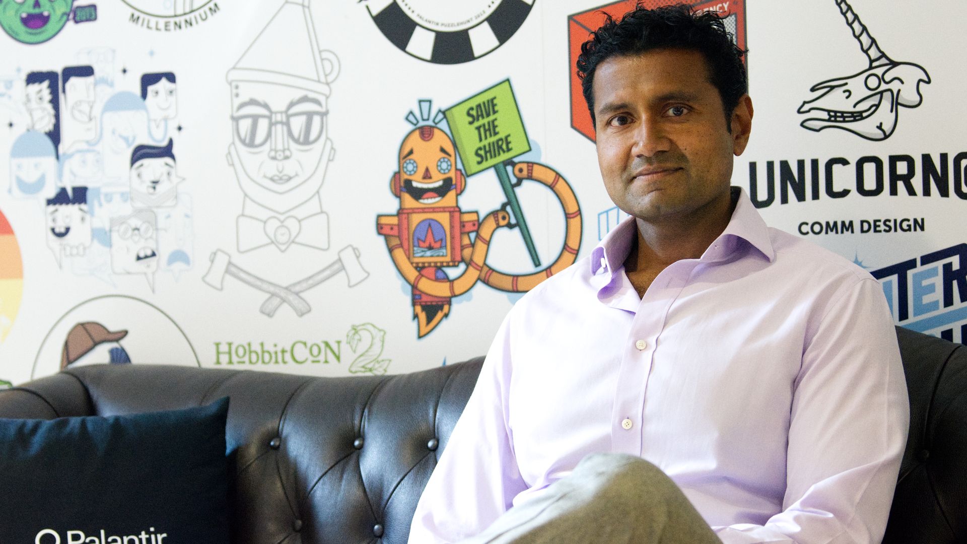 Shyam Sankar is seated on a brown leather couch. He is wearing a pink dress shirt and khaki pants. The background is full of colorful logos.