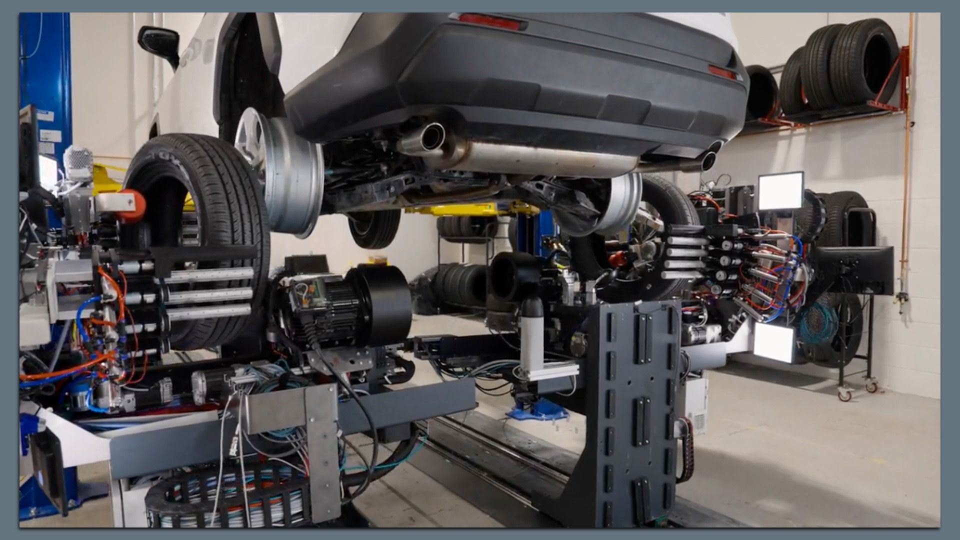 White car on a lift in a busy tire service shop, where a robot from Automated Tire rotates and balances the tires. In the background stacked tires are visible in a workshop.