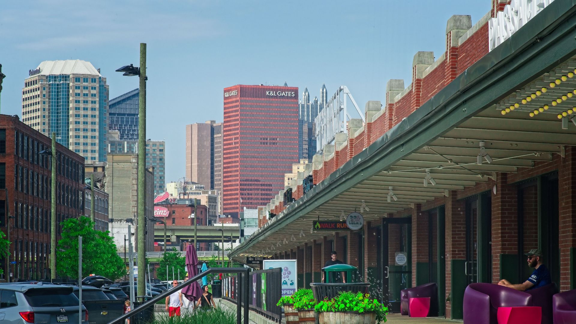 Street view with parked cars, greenery, people walking, colorful outdoor seating, and skyscrapers including a red K&L Gates building under a clear blue sky in Pittsburgh, Pa.