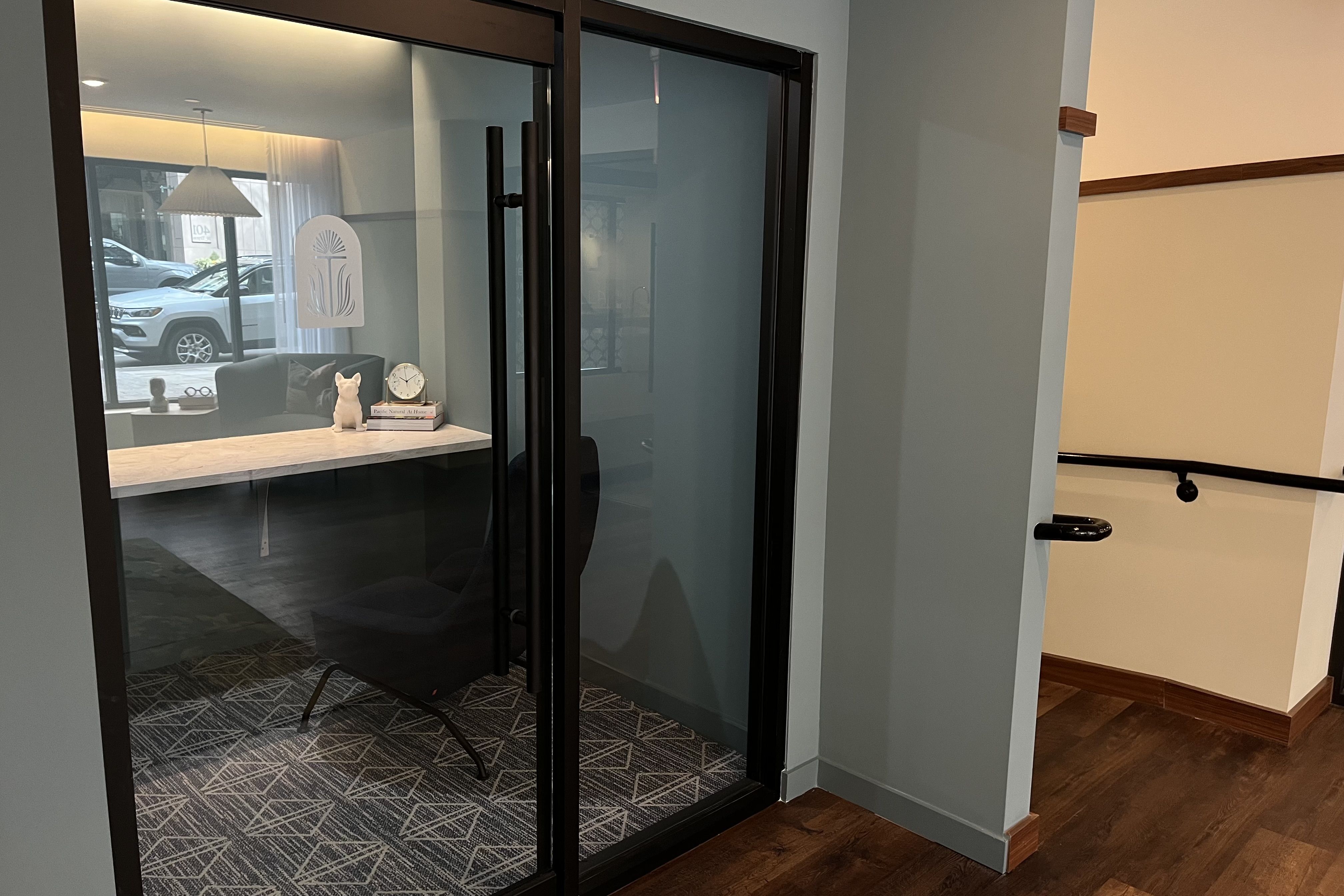 Glass-walled office with black chair and patterned carpet, white clock and dog figurine on marble counter, and view of street with parked cars outside window.