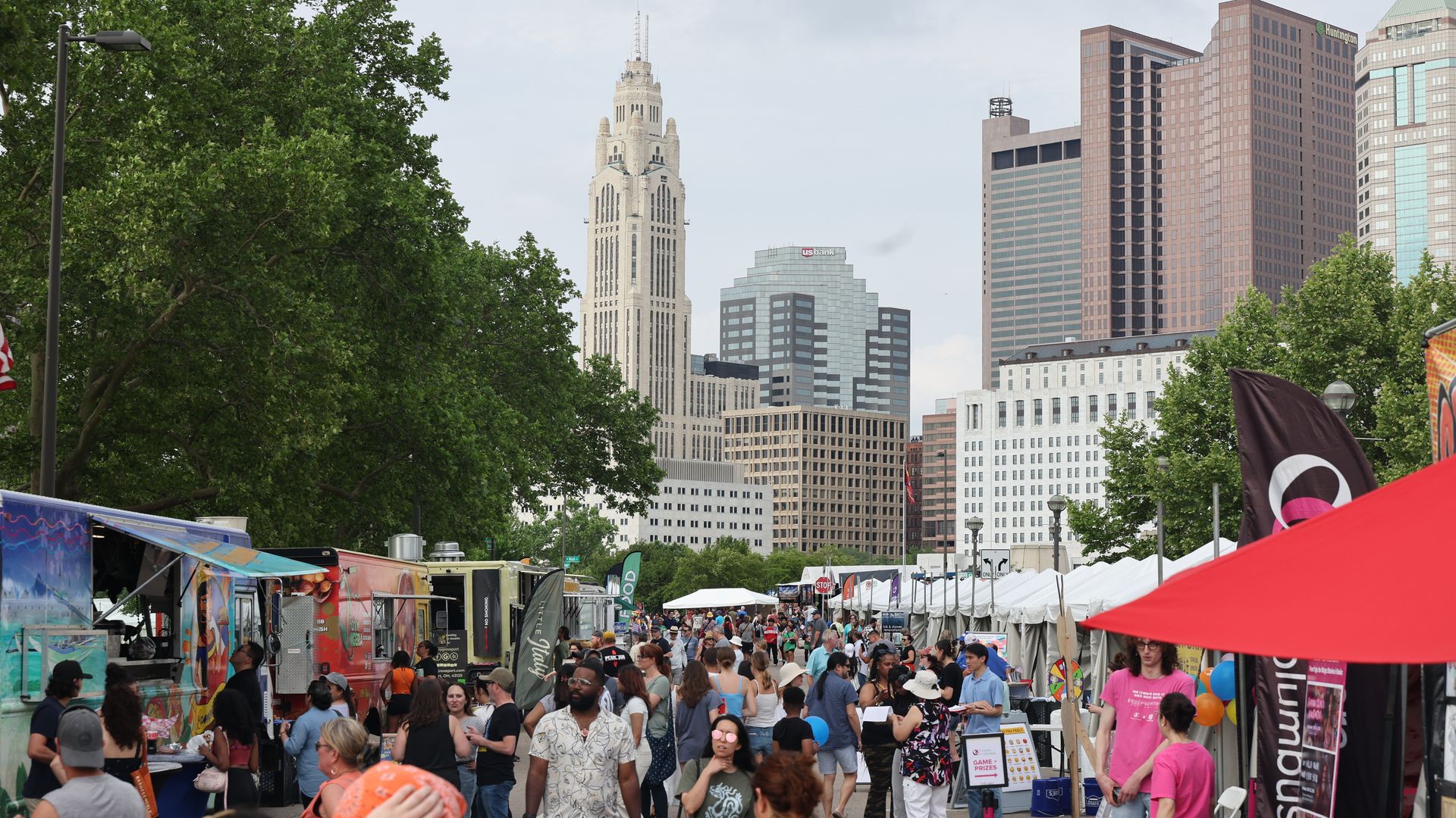 Busy city street festival with food trucks and white tents, crowded with people. In the background, a tall ornate white tower rises between modern office buildings, framed by green trees.