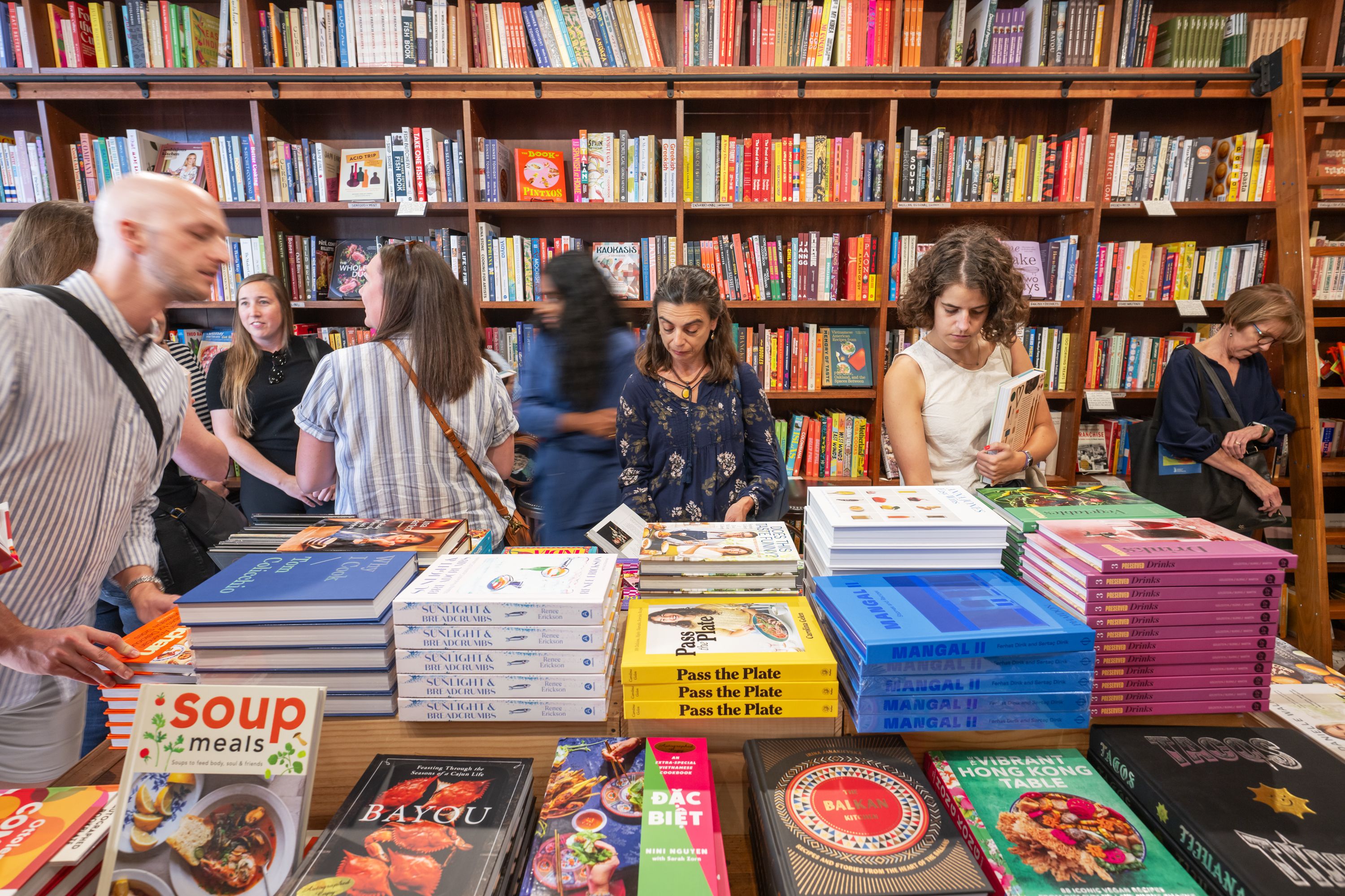 Busy bookstore with tall wooden shelves of colorful books; several shoppers browse tables and stacks of cookbooks and magazines in a bright, crowded shop.