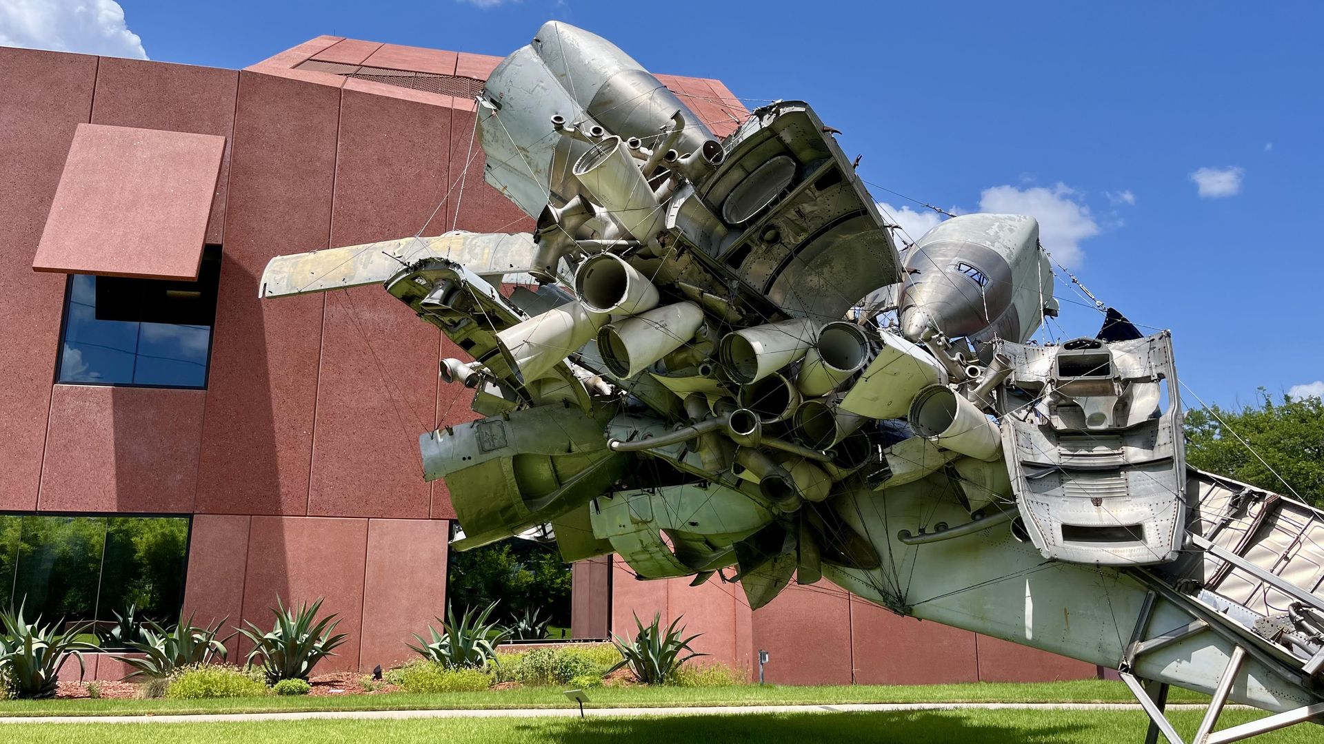 A metal sculpture sits above a grassy yard in front of a bright red building with clean lines and square windows.