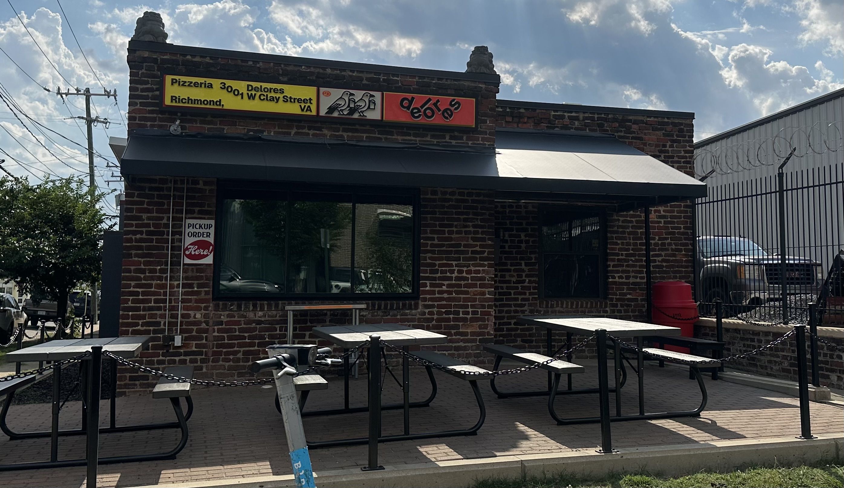 Brick pizzeria with a yellow sign reading Pizzeria 3001 W Clay Street Richmond VA and a red logo. Outdoor metal tables under a gray awning; a barbed-wire fence and a parked SUV in daylight.