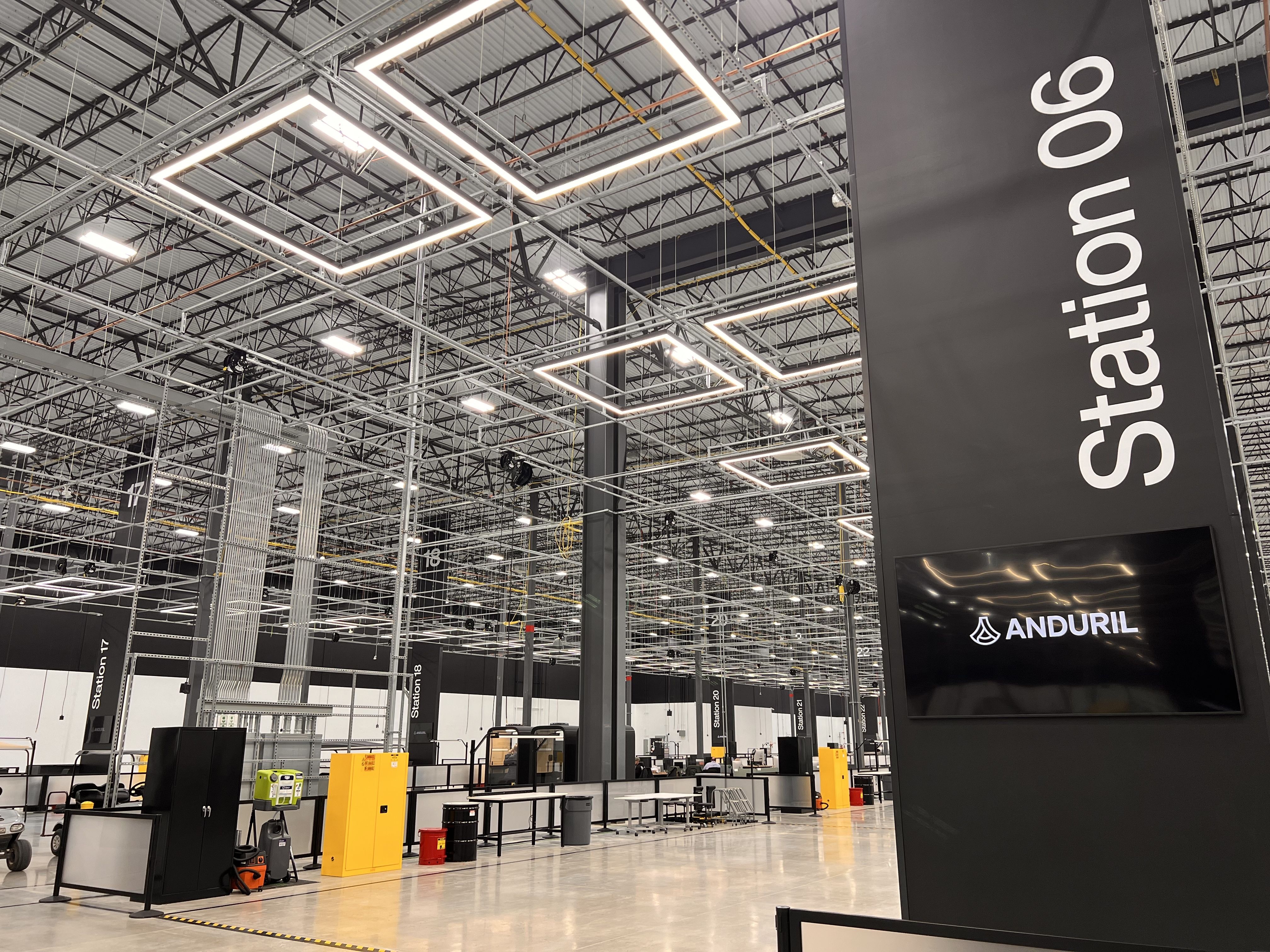 Spacious warehouse-style exhibition hall with an exposed metal ceiling and geometric square LED lights. A tall black pillar on the right displays "Station 06" and the ANDURIL logo on a screen; yellow cabinets and equipment line the floor.