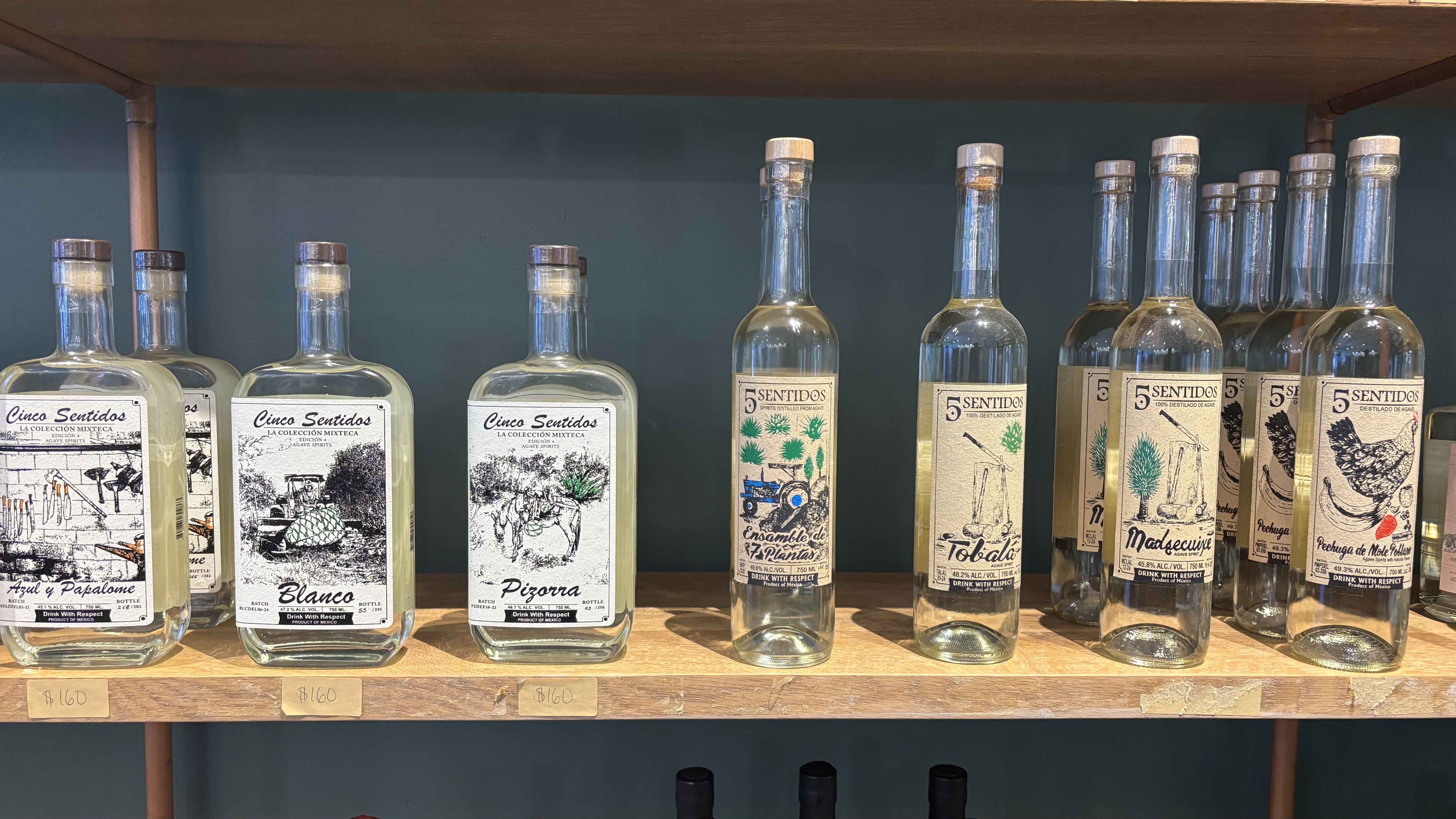 Clear glass bottles with wooden corks displayed on wooden shelves, labeled 'Cinco Sentidos' and '5 Sentidos' with black and colored illustrations, priced at 160 pesos.