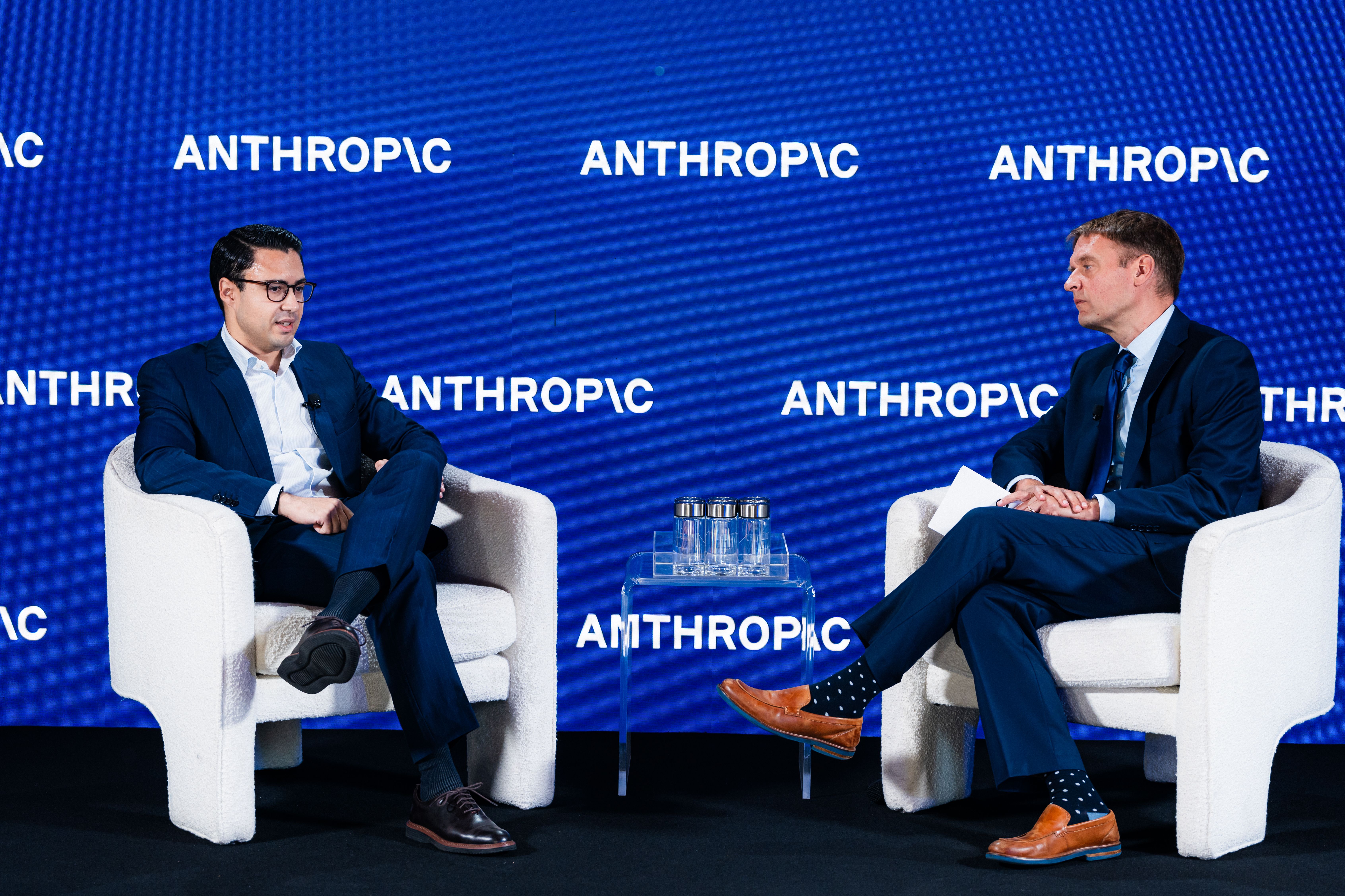 Two men in dark suits sit on white chairs on a blue stage backdrop that repeats "ANTHROPIC"; a small glass table with water bottles sits between them as they converse.
