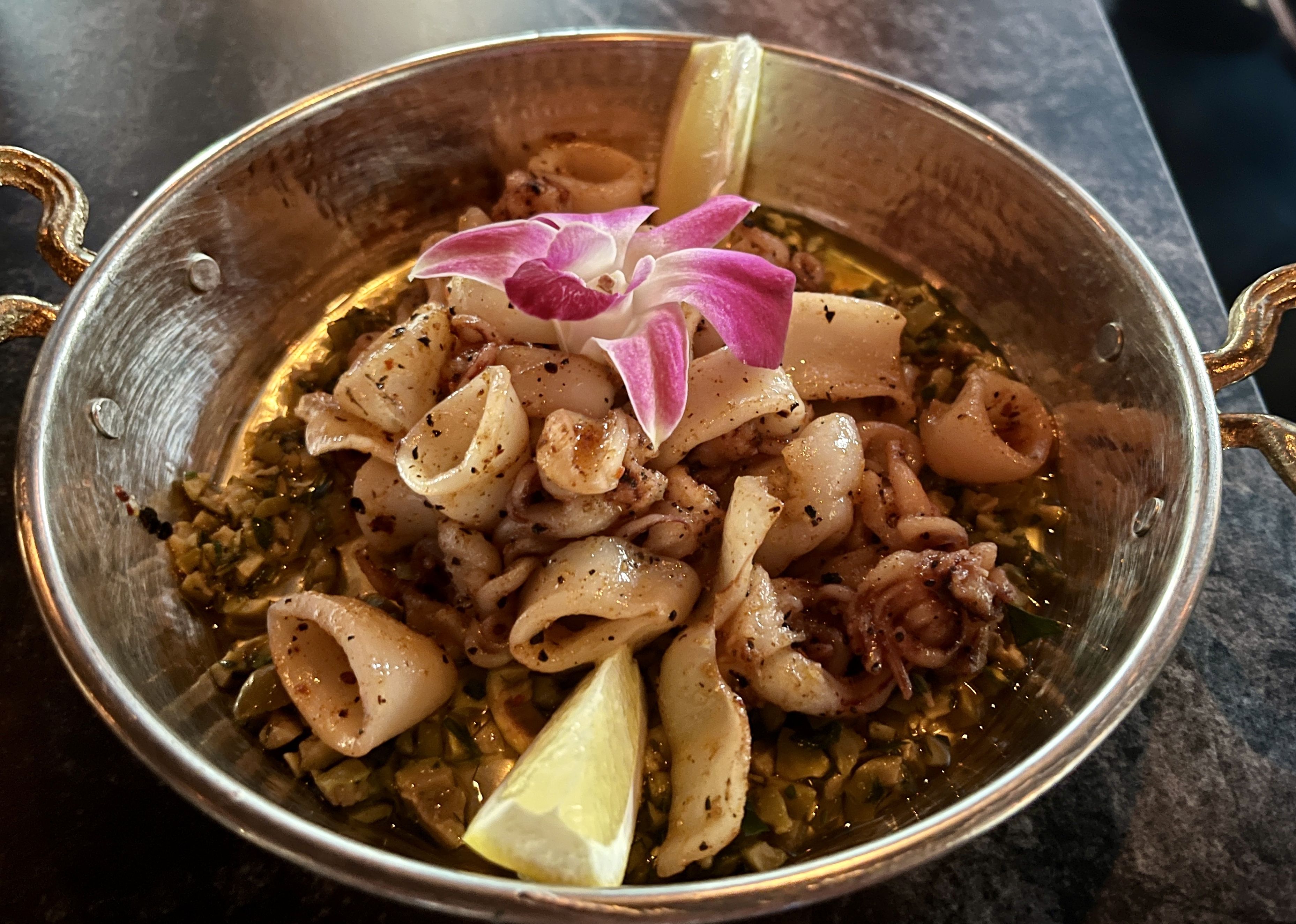 Metal bowl with grilled squid rings and tentacles, garnished with a pink orchid flower and two lemon wedges, resting on a bed of chopped green herbs and oil.
