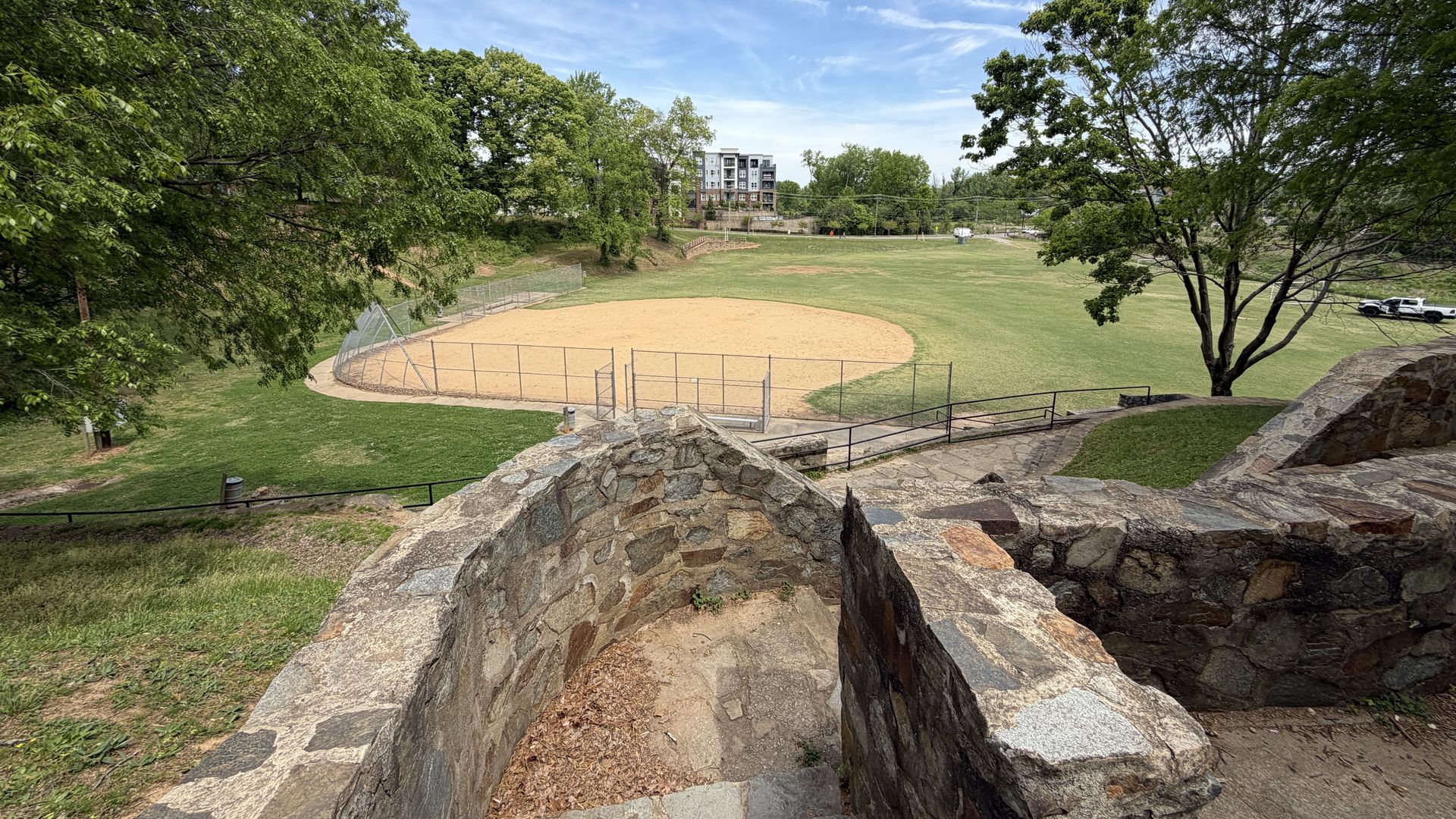 View from a stone terrace over a baseball field: sandy infield, green outfield, chain-link fencing, and tall trees. A distant building and parked cars appear beyond the park.