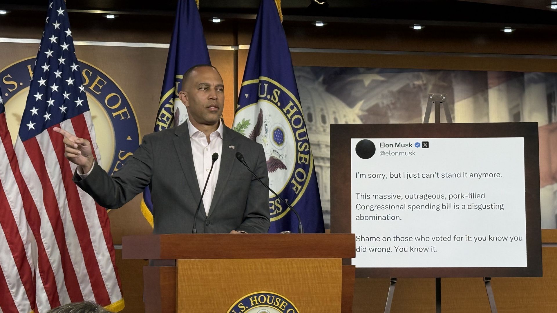 House Minority Leader Hakeem Jeffries holds up a finger in front of an American flag behind a wooden podium next to a blown-up graphic of an Elon Musk tweet.