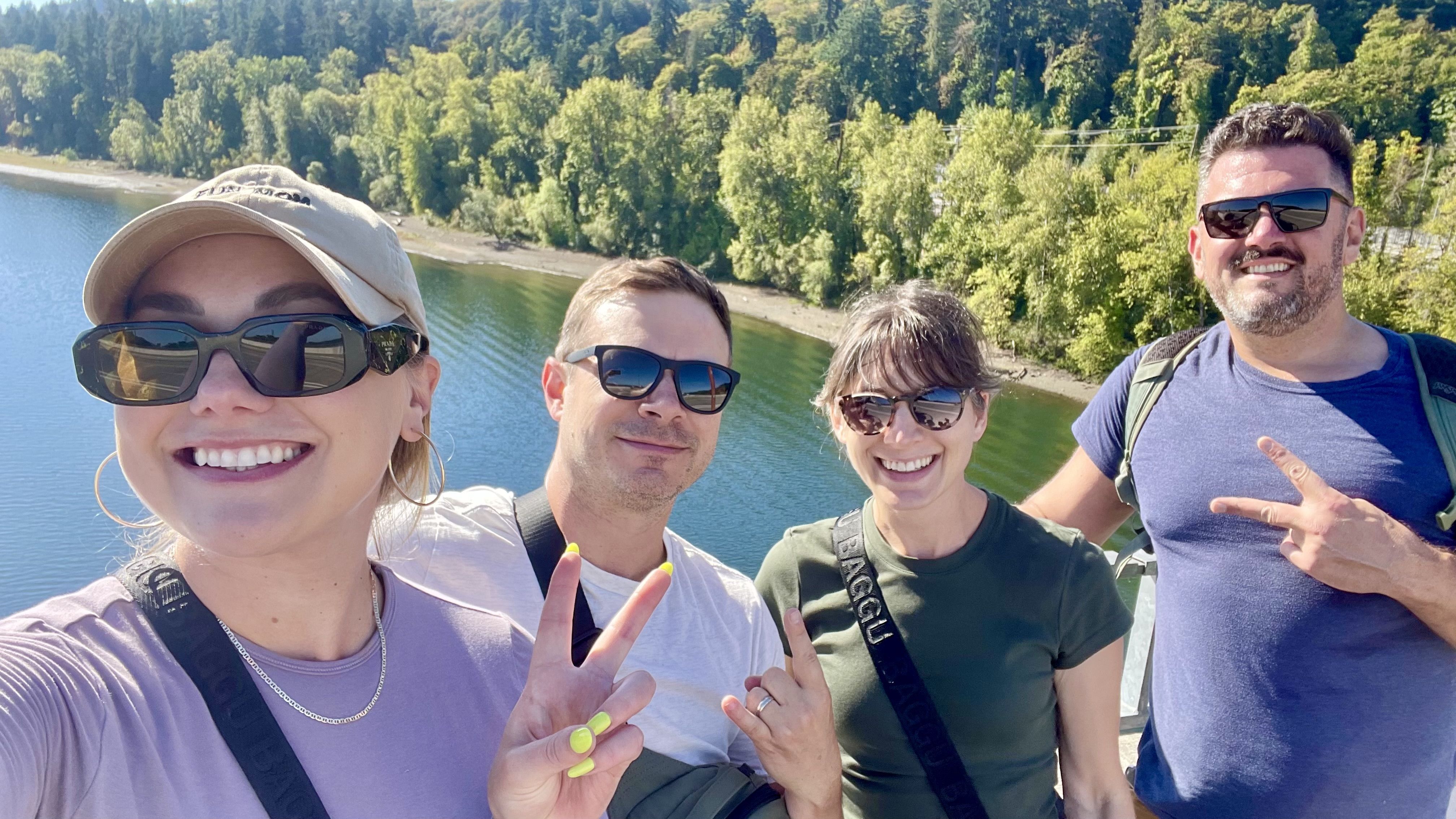 Four friends on a sunny boat ride posing for a selfie: woman in beige cap front left, man in white shirt, woman with bangs in green, and bearded man in blue shirt; lake and forest behind.