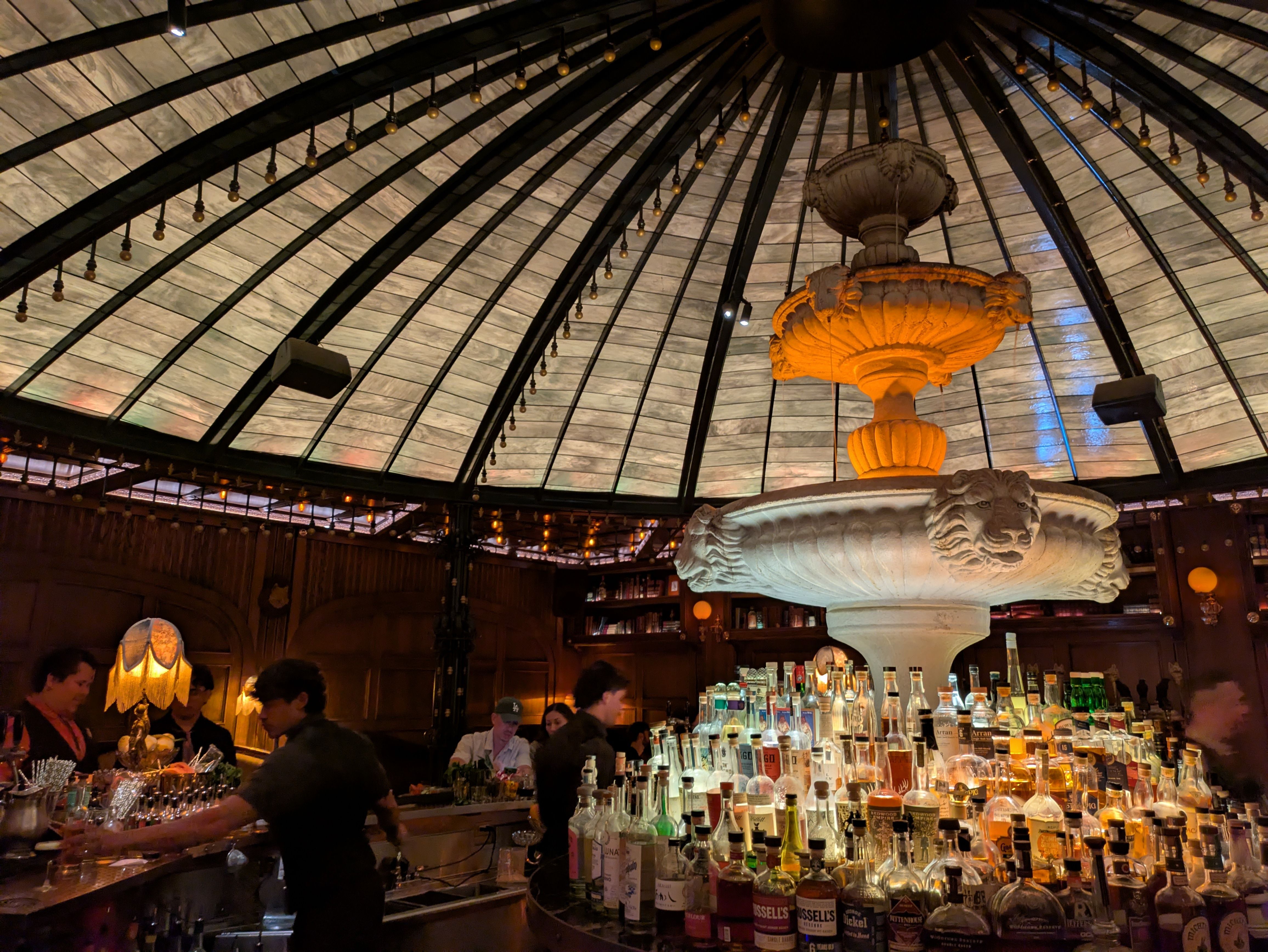 Dim bar interior with vaulted glass ceiling and dark beams; a large ornate orange chandelier above a long counter filled with bottles, as patrons and bartenders work.