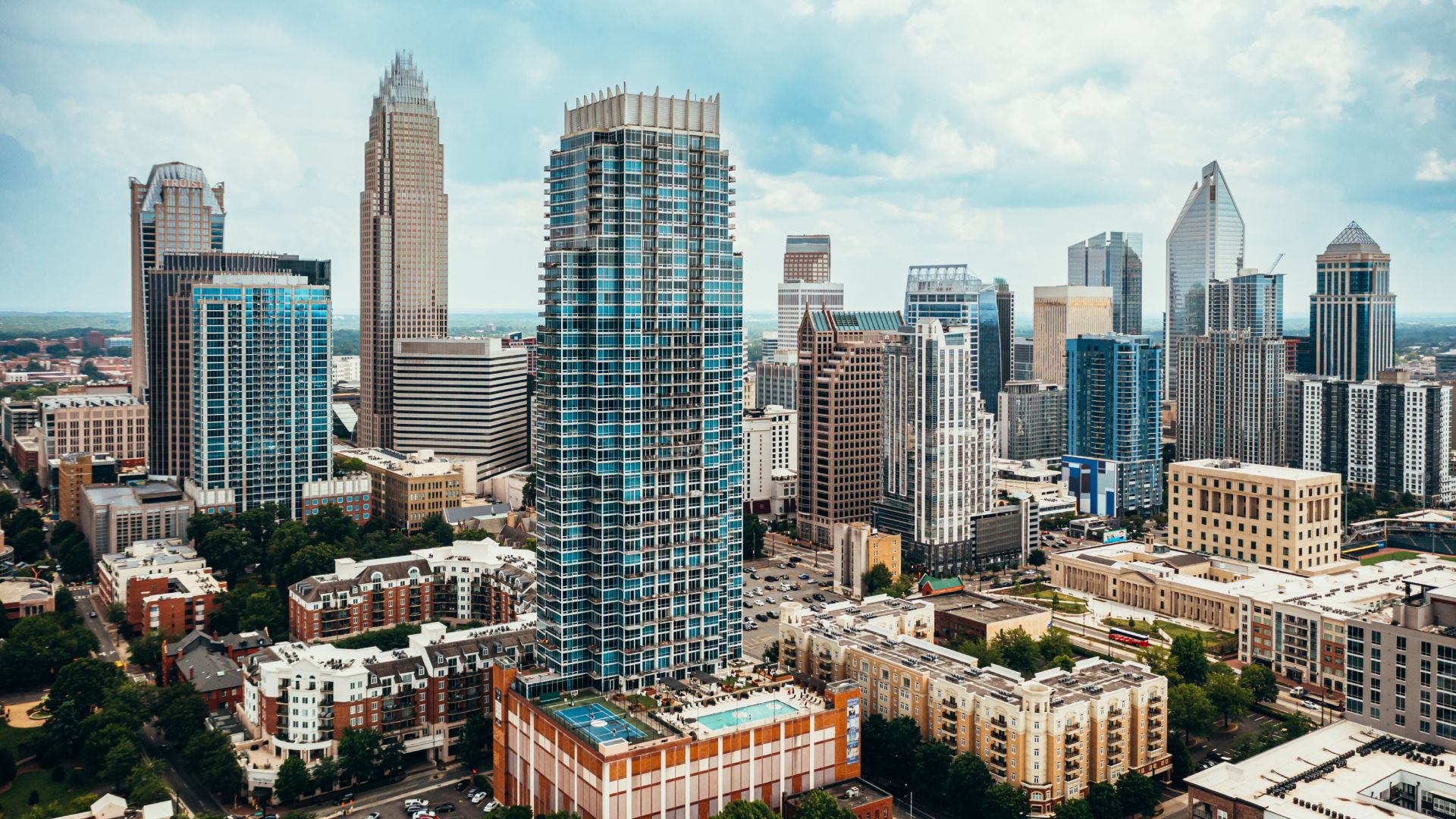 An aerial city skyline featuring a cluster of glass skyscrapers in blue and gray, surrounded by mid and low-rise buildings, streets, trees and a bright, cloudy sky.