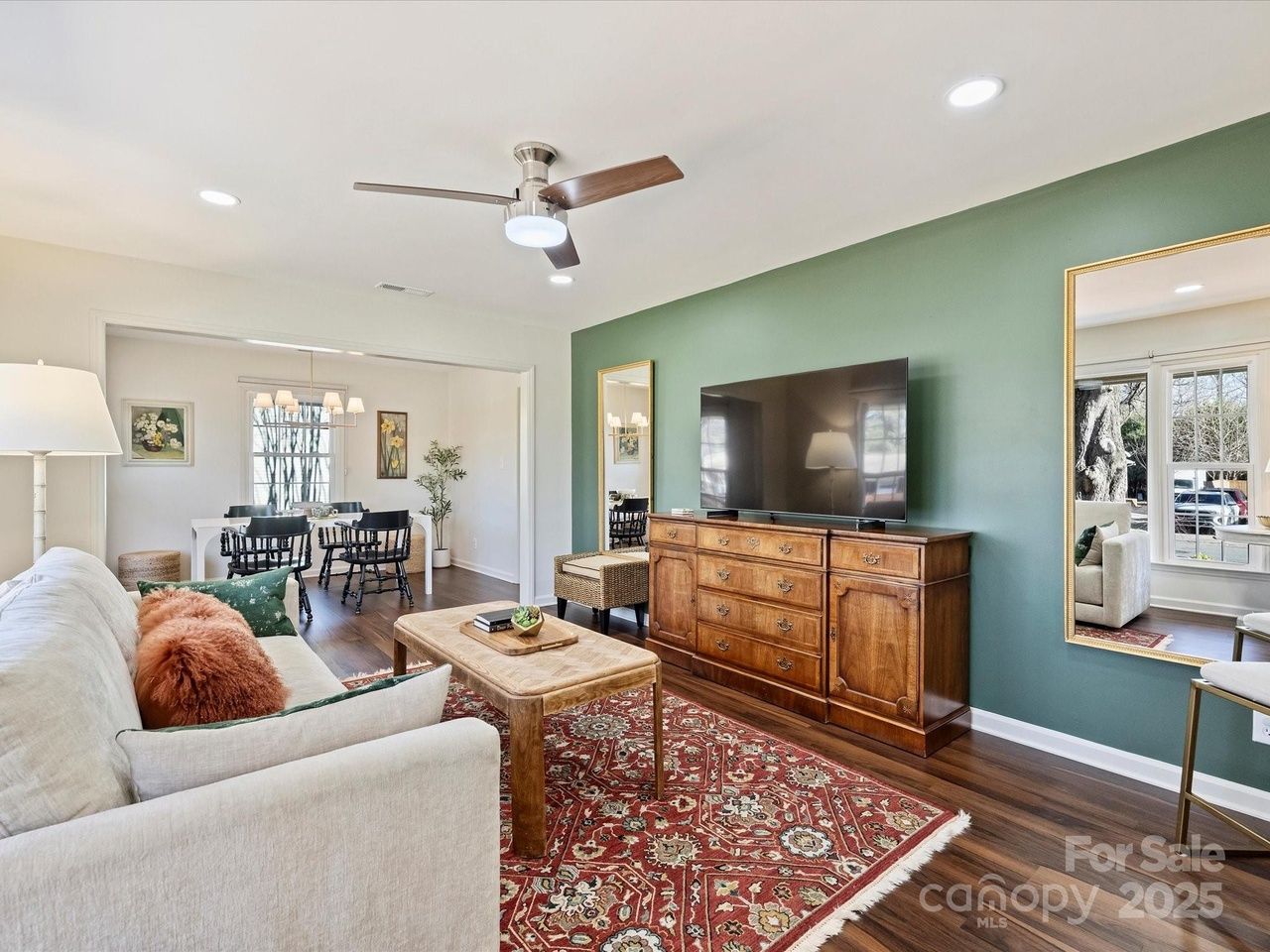 Bright living room with beige sofa and orange pillow, wooden coffee table on red patterned rug, green accent wall with large mirror and TV on wooden dresser, visible dining area beyond.
