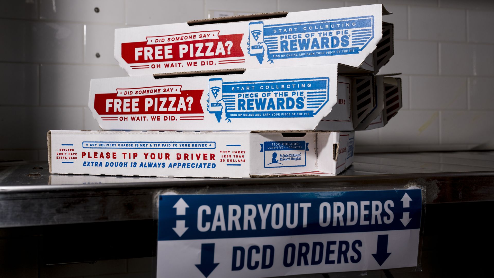 Stacked white pizza boxes with red 'FREE PIZZA?' and blue 'REWARDS' panels, on a shelf in a kitchen; a blue 'CARRYOUT ORDERS' sign is visible below.