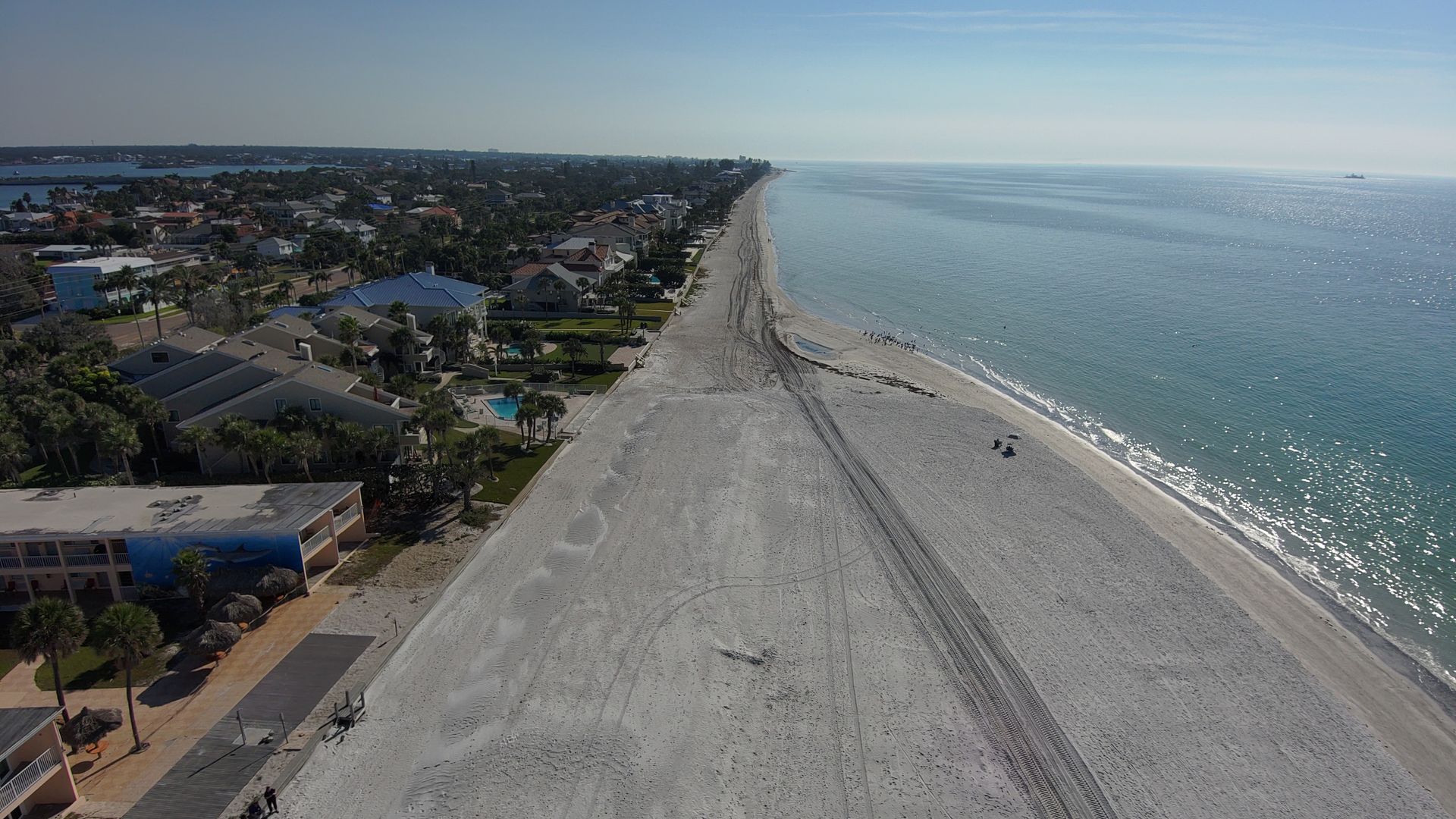 Aerial view of a coastal town with a long sandy beach along the sea, rows of houses and palm trees on the left, a road along the shore, and tire tracks in the sand under a sky.