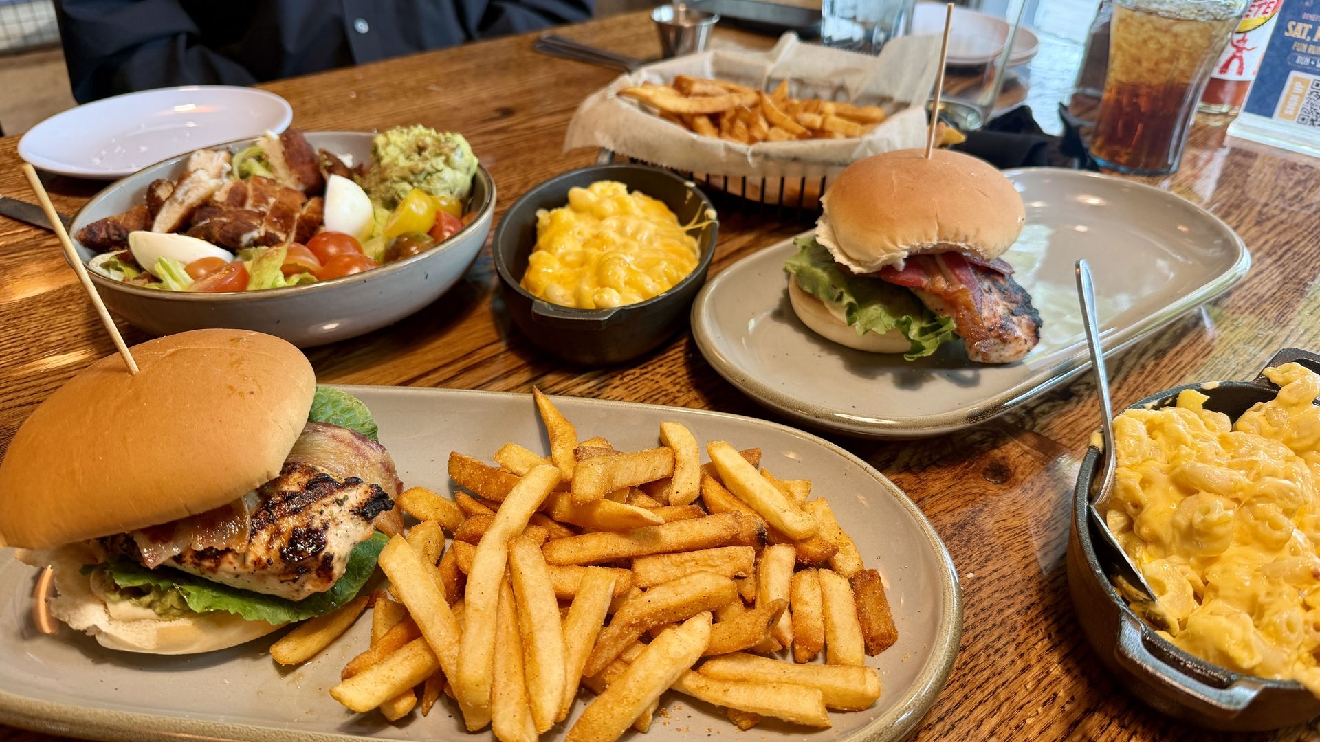 Top-down view of a wooden table with a bacon cheeseburger and fries, a salad bowl with eggs, tomatoes, and avocado, a macaroni and cheese dish, and a basket of fries; drinks and a person in a dark shirt are in the background.