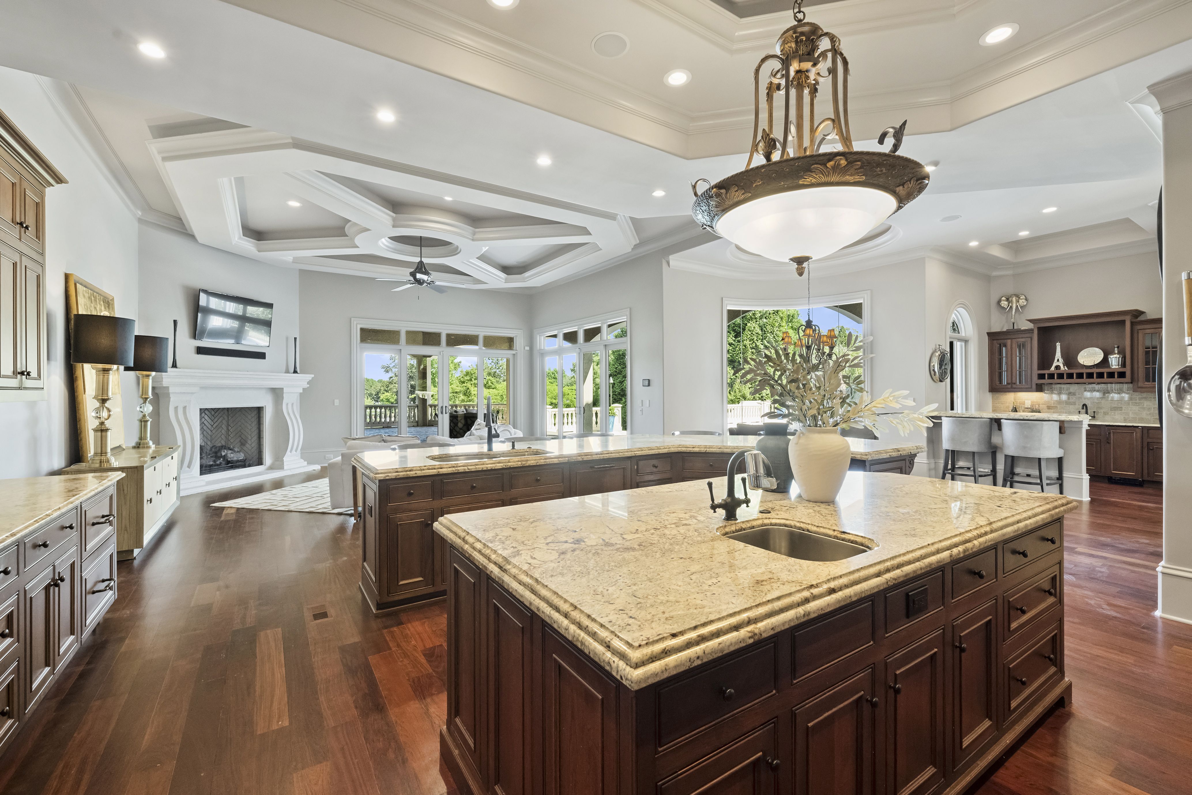 Bright open kitchen with dark wood cabinets and beige granite island on hardwood floors. Coffered ceiling, ornate chandelier, and recessed lights; adjacent living area with a fireplace, TV, and large glass doors.