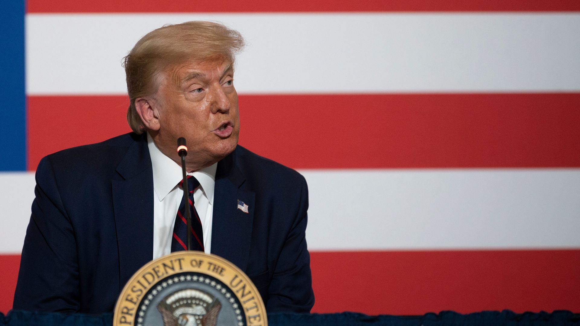 Donald Trump speaking at a podium with a large American flag in the background 