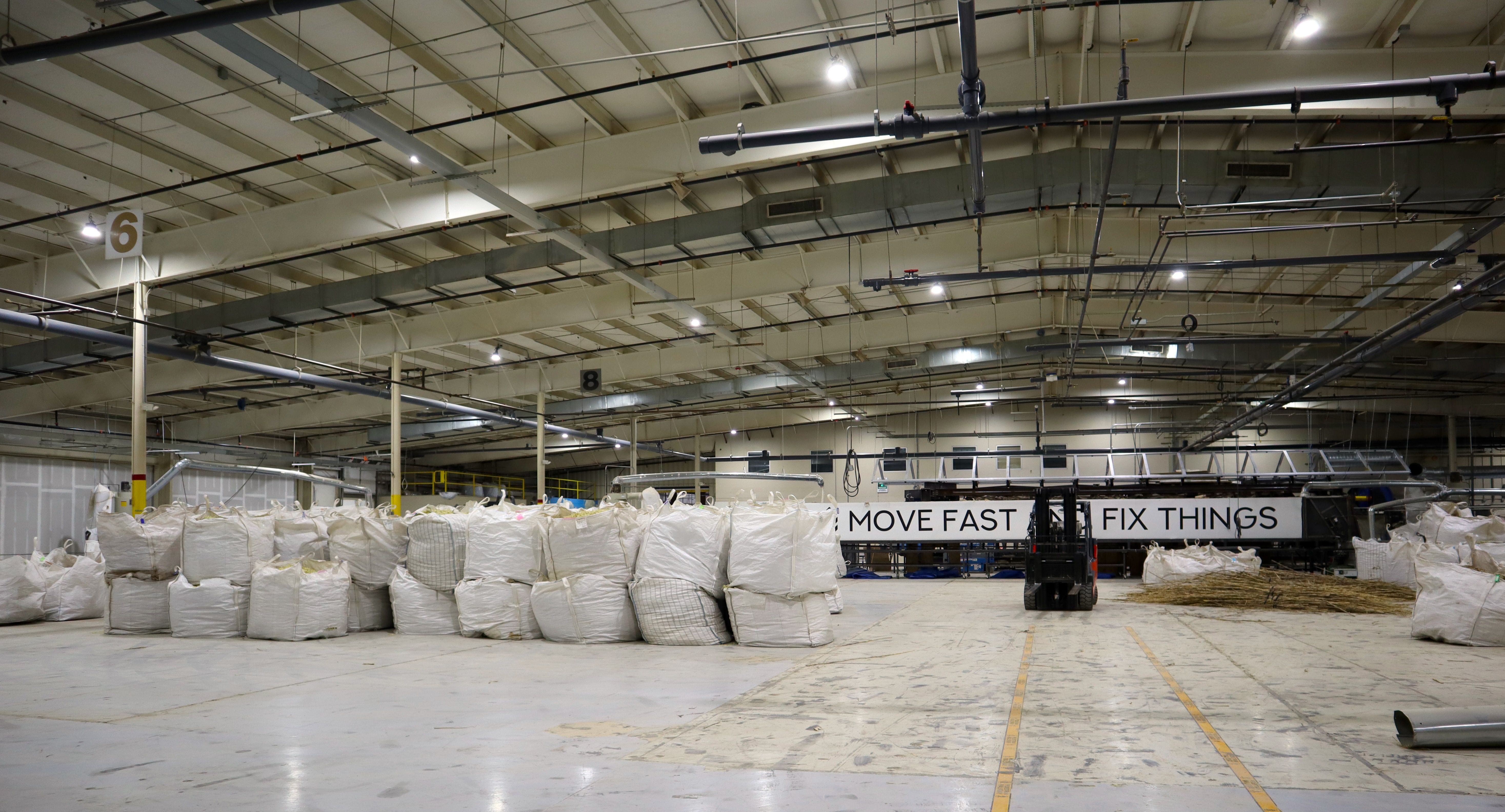 Inside a large warehouse with overhead pipes, white bulk bags lined up along the floor. A forklift stands near a long banner that reads "MOVE FAST FIX THINGS".