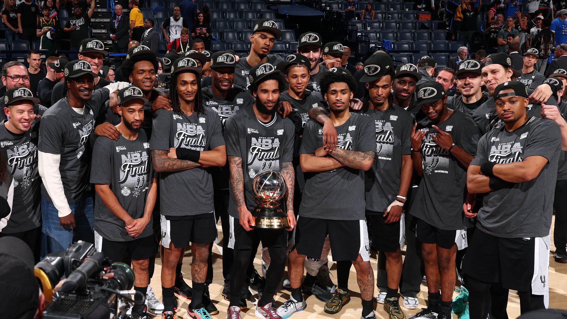 Group of basketball players wearing grey Finals shirts and caps pose for a team photo around the NBA Finals trophy on the court, with fans, photographers, and arena seating in the background.