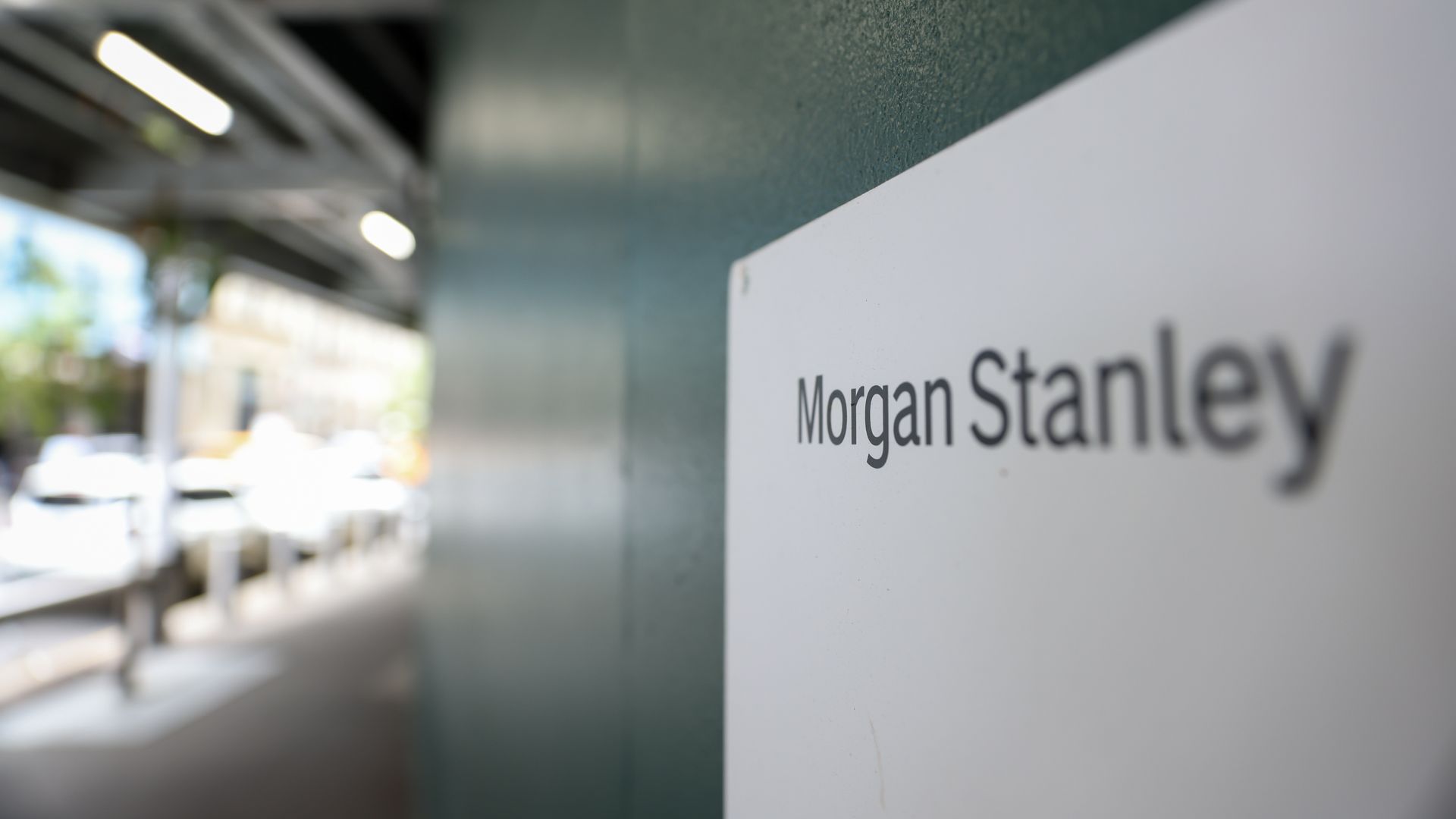 Close-up of a white sign reading "Morgan Stanley" in black, mounted on a teal wall; blurred interior with bright overhead lights in the background.